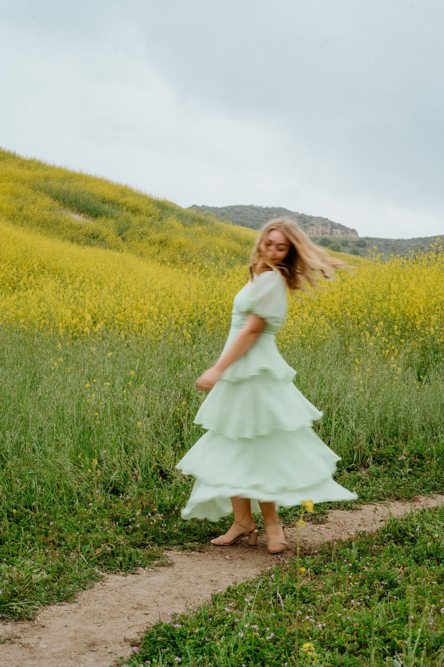 Woman in mint tiered dress spinning on dirt path in yellow flower field.