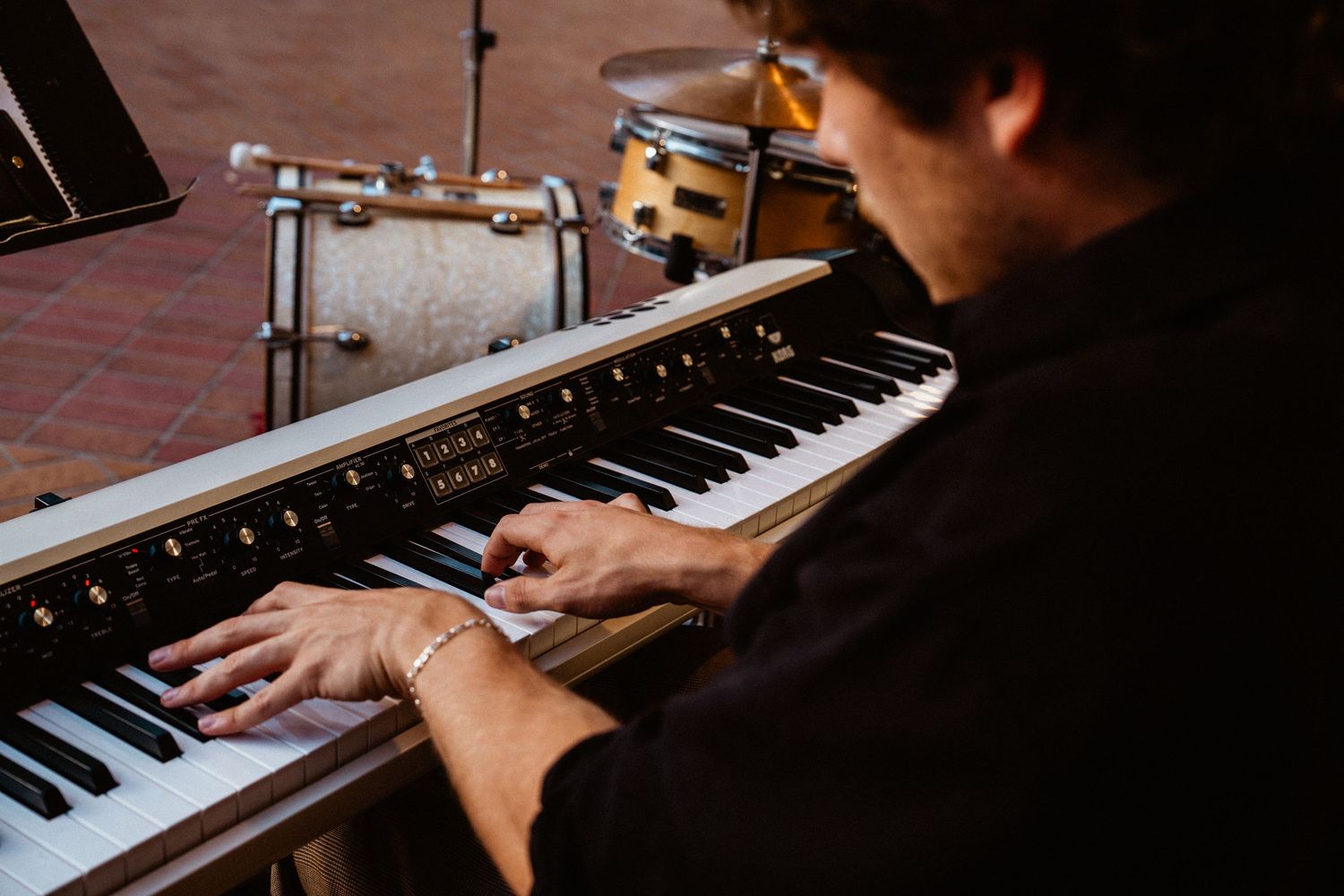 Musician playing a keyboard piano at an outdoor event with a drum kit visible in the background.