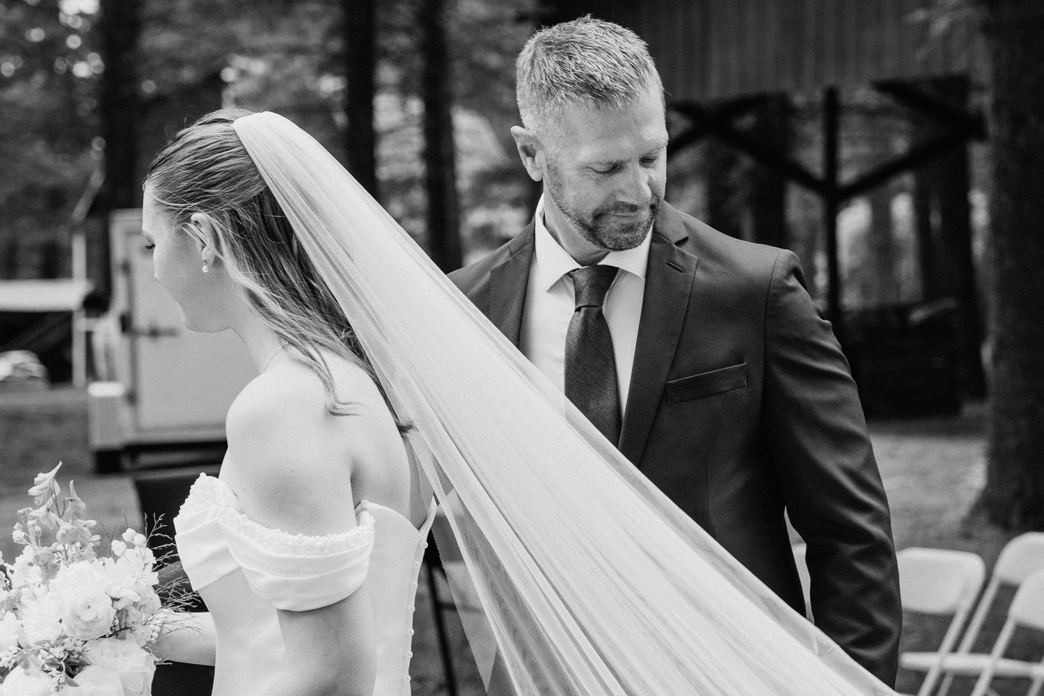 Black and white photo of groom adjusting bride's veil outdoors at a woodland wedding ceremony.