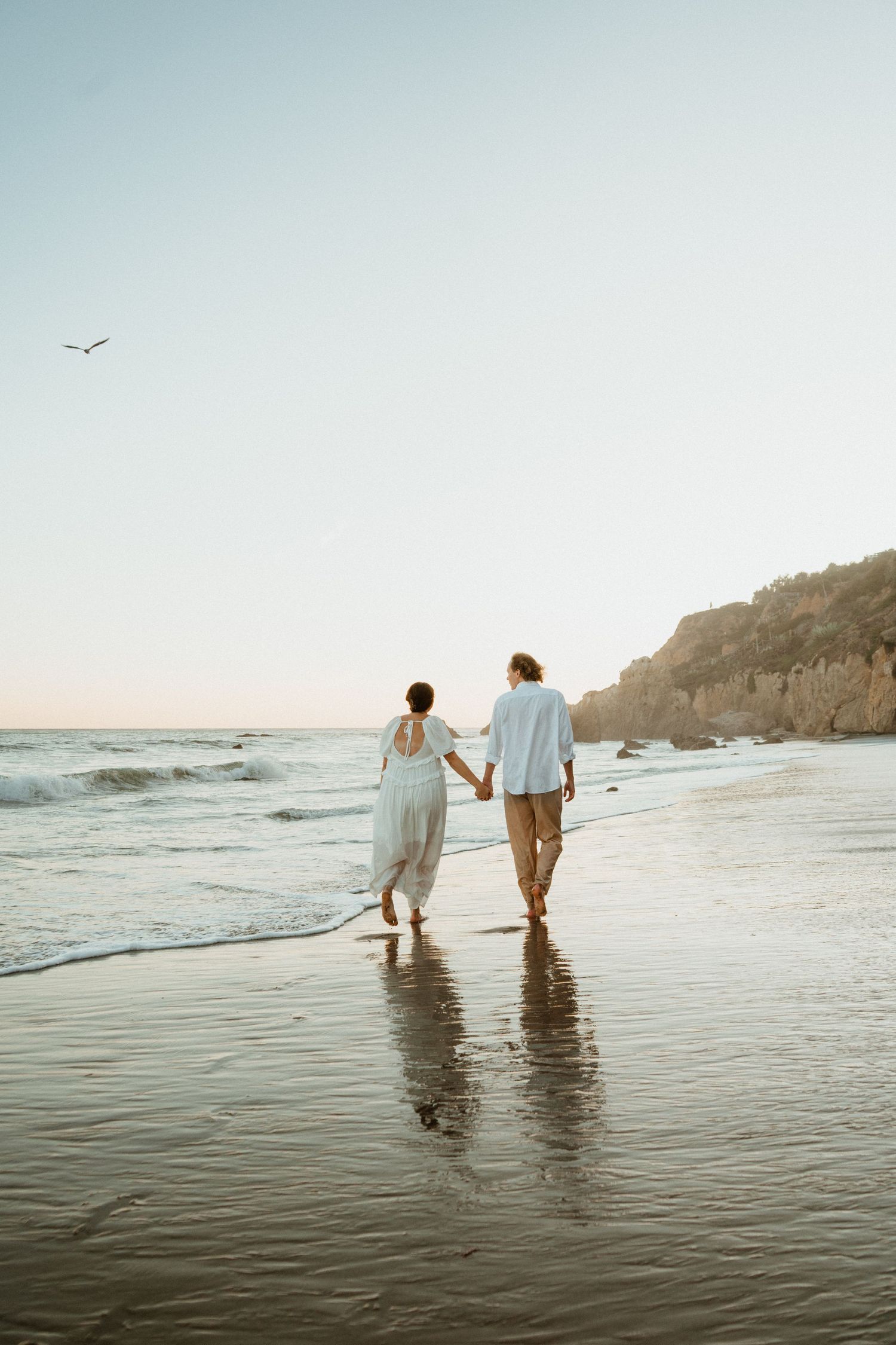 A couple walks hand-in-hand along a serene beach at sunset, wearing white outfits with ocean waves beside them.