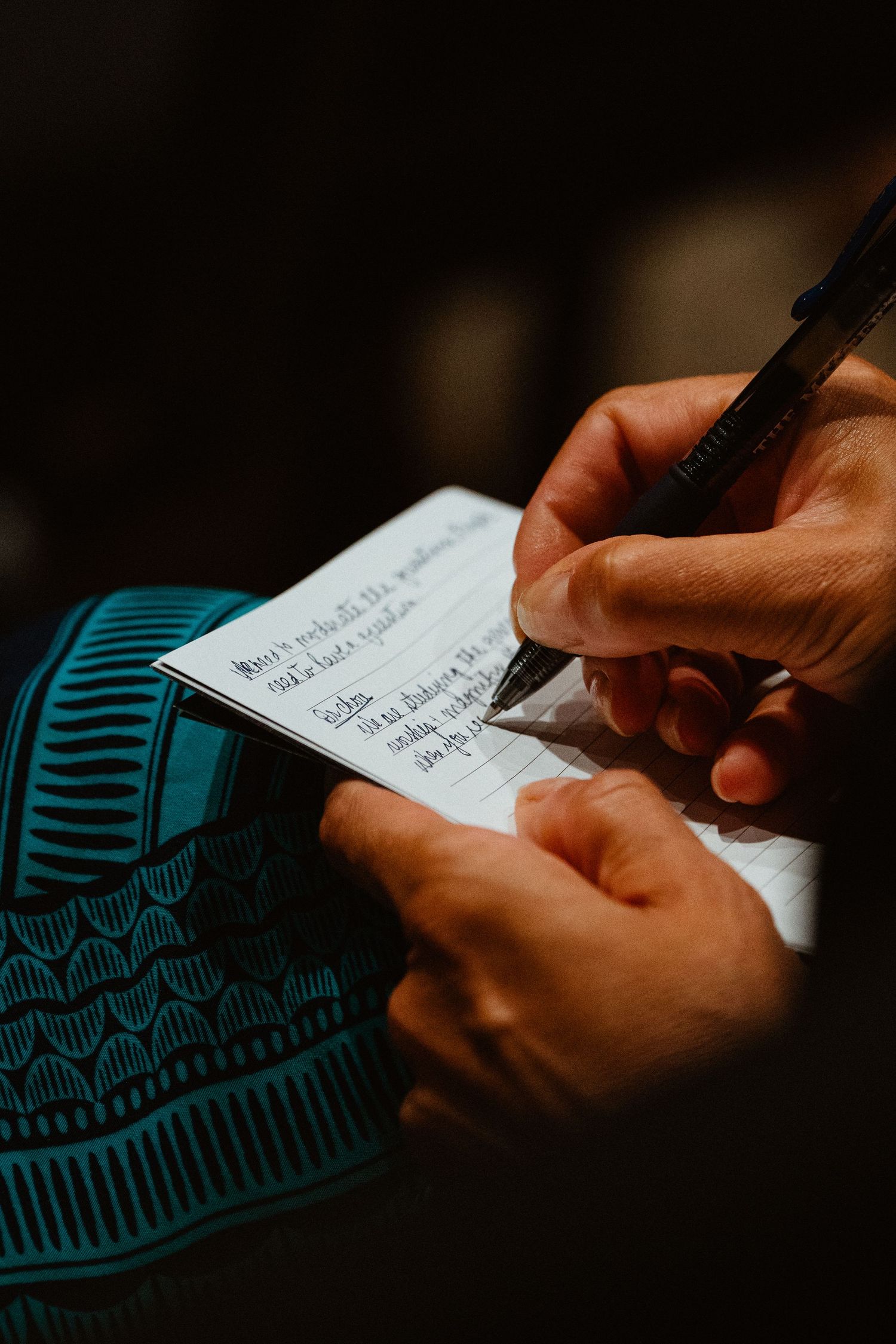 Close-up of hands writing notes on a small card with a pen, wearing a teal patterned sleeve.