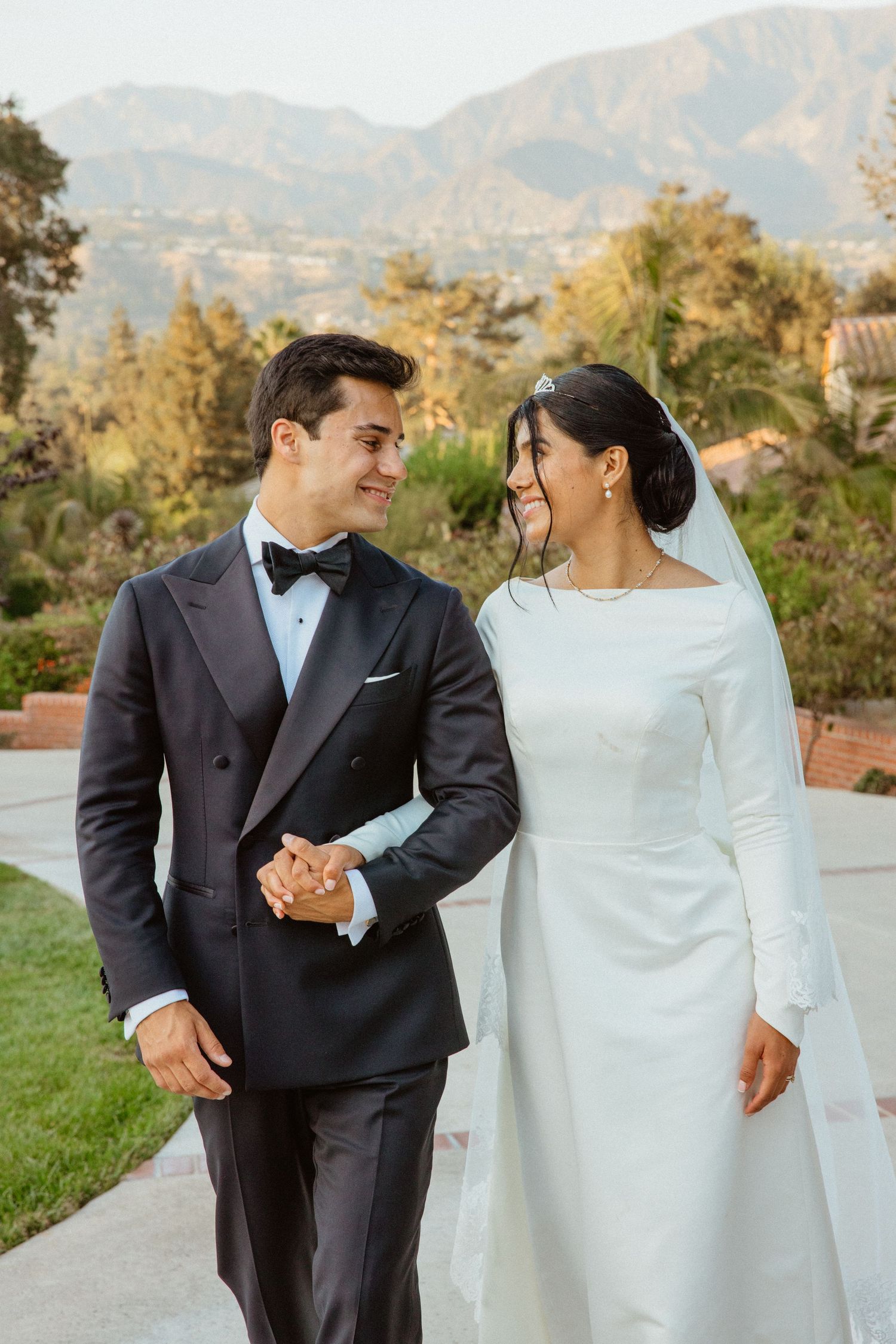 Bride in long-sleeve white gown and groom in black tuxedo walk hand in hand with mountain backdrop.