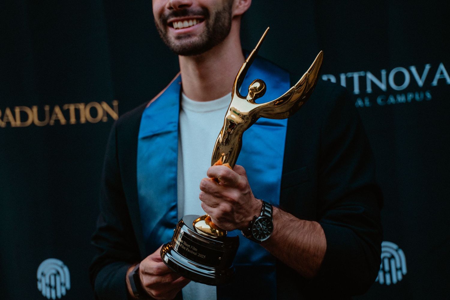 Smiling man in graduation attire holding a gold trophy award at a ceremony.