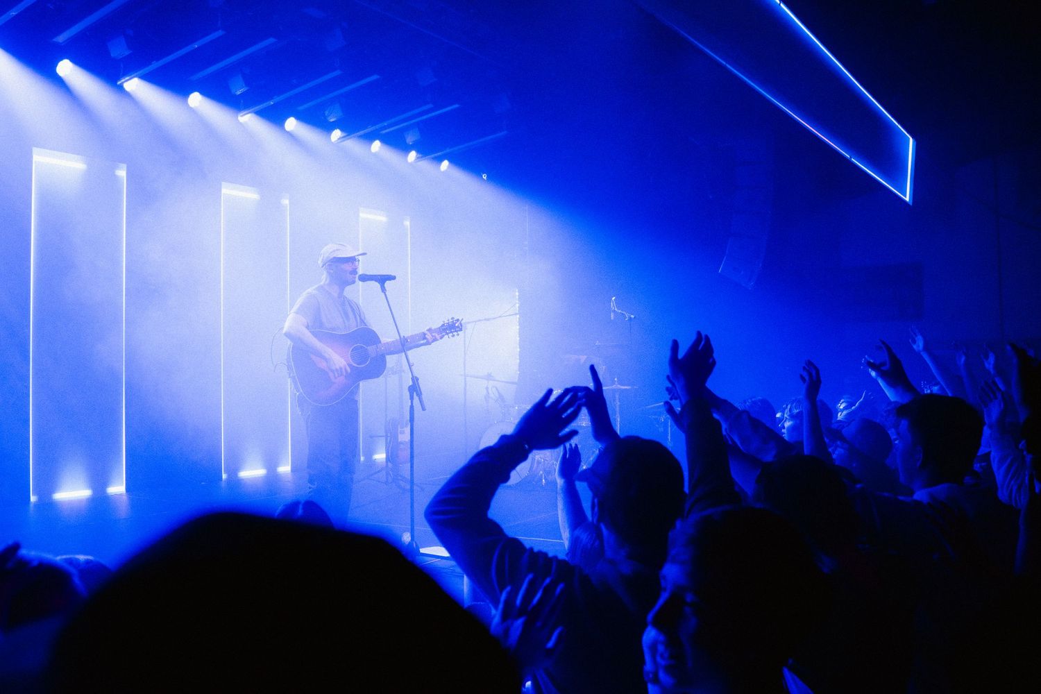 Musician performing on stage with blue lighting and fog at a live concert with crowd raising hands.