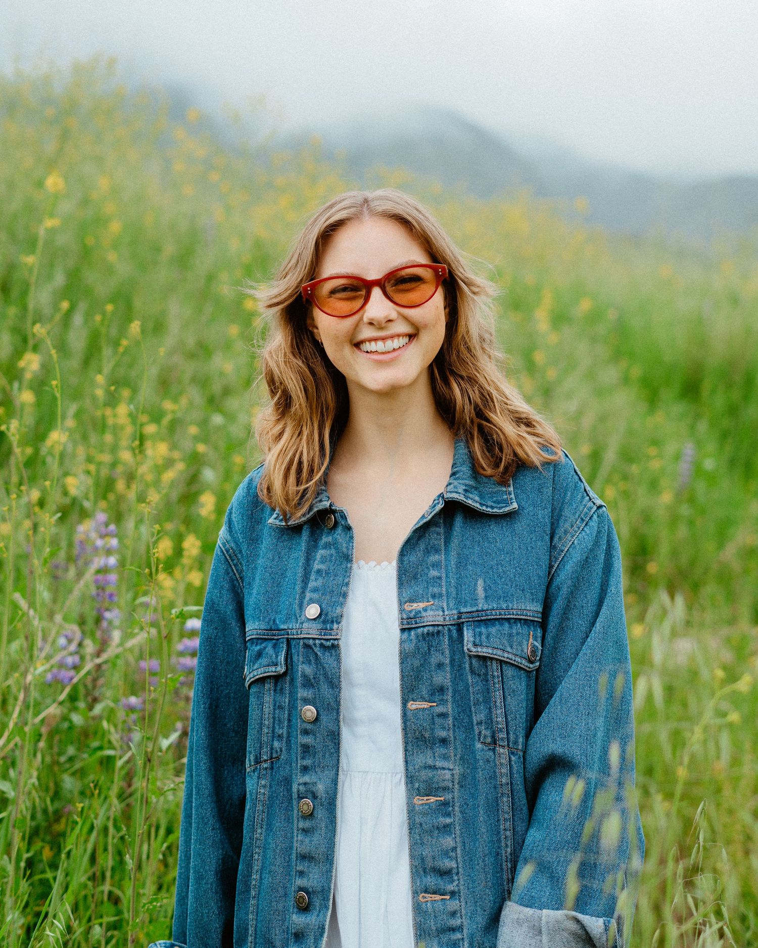Smiling young woman in denim jacket and orange sunglasses stands in colorful wildflower meadow.