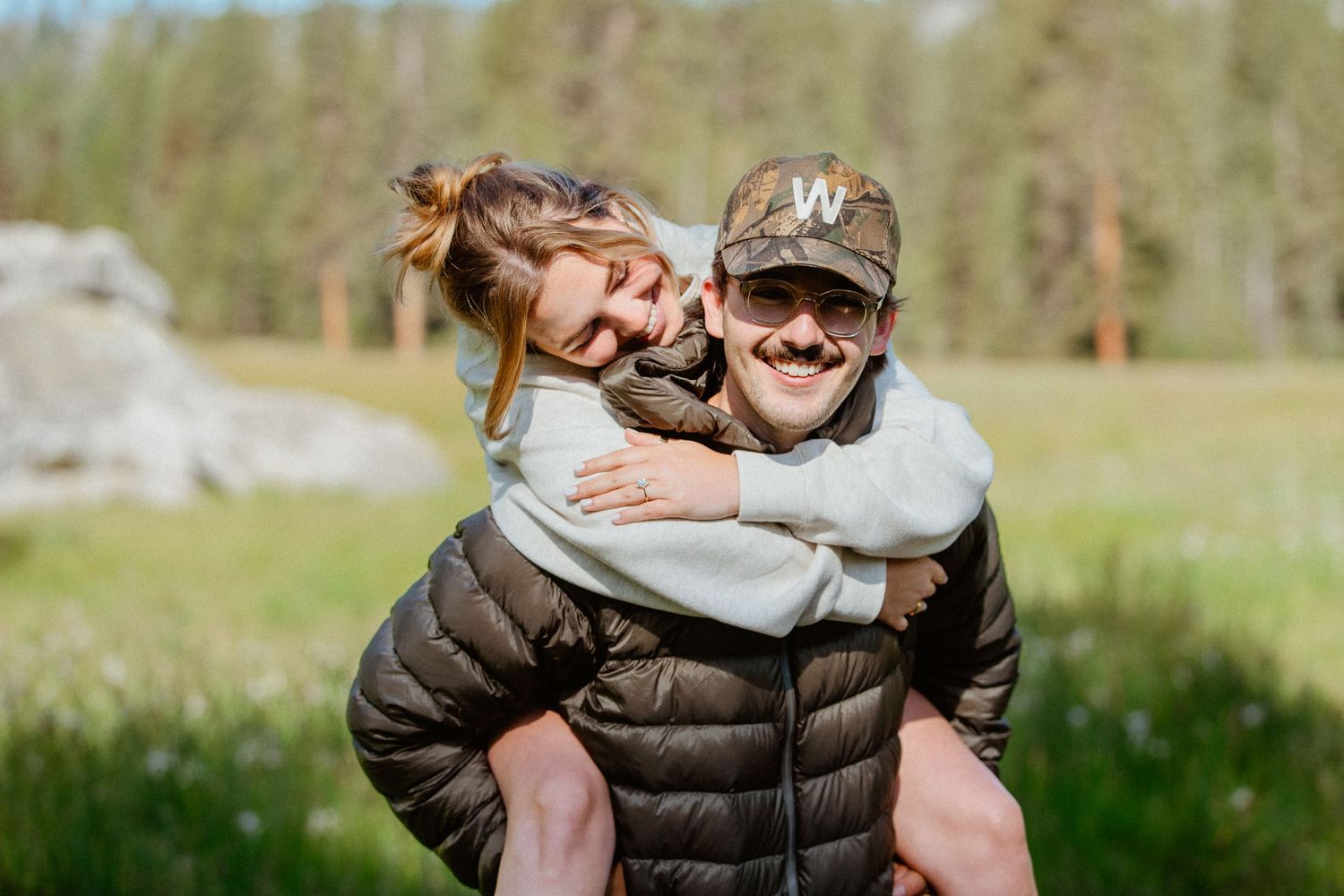 Couple laughing outdoors, man giving woman a piggyback ride in a scenic green field.