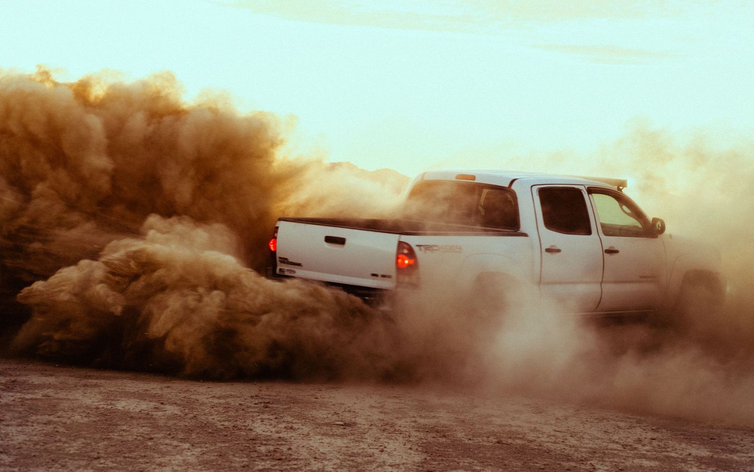 White pickup truck surrounded by billowing brown dust cloud while off-roading in desert.