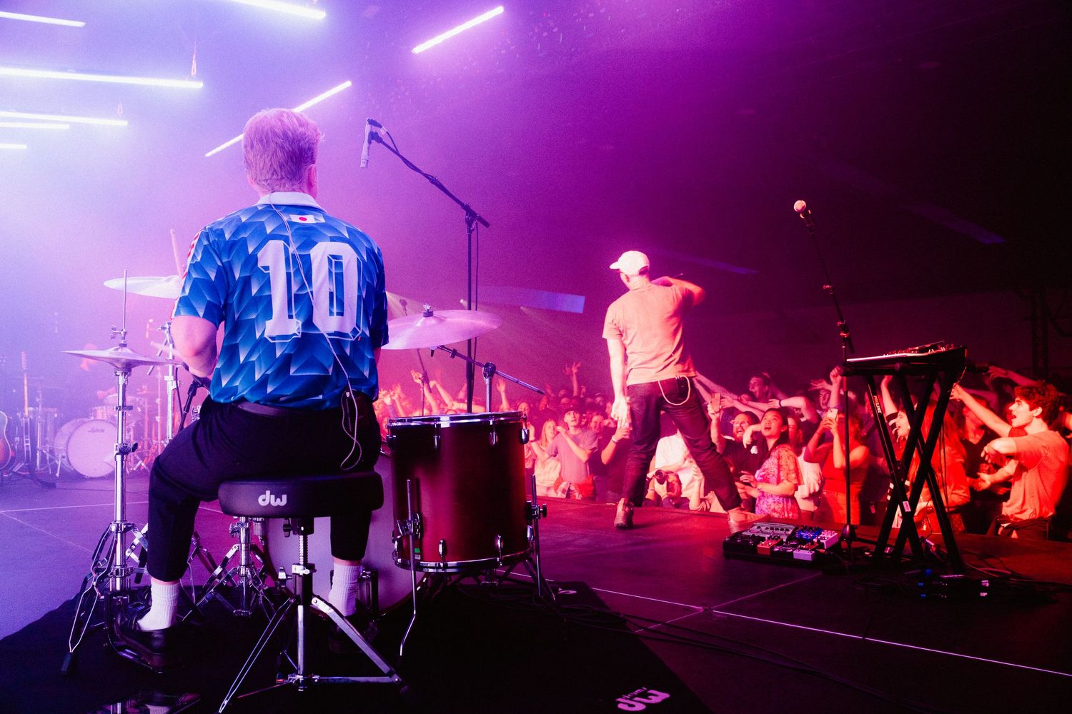 A drummer wearing a blue number 10 jersey plays as the lead singer performs in front of the crowd, purple lighting.