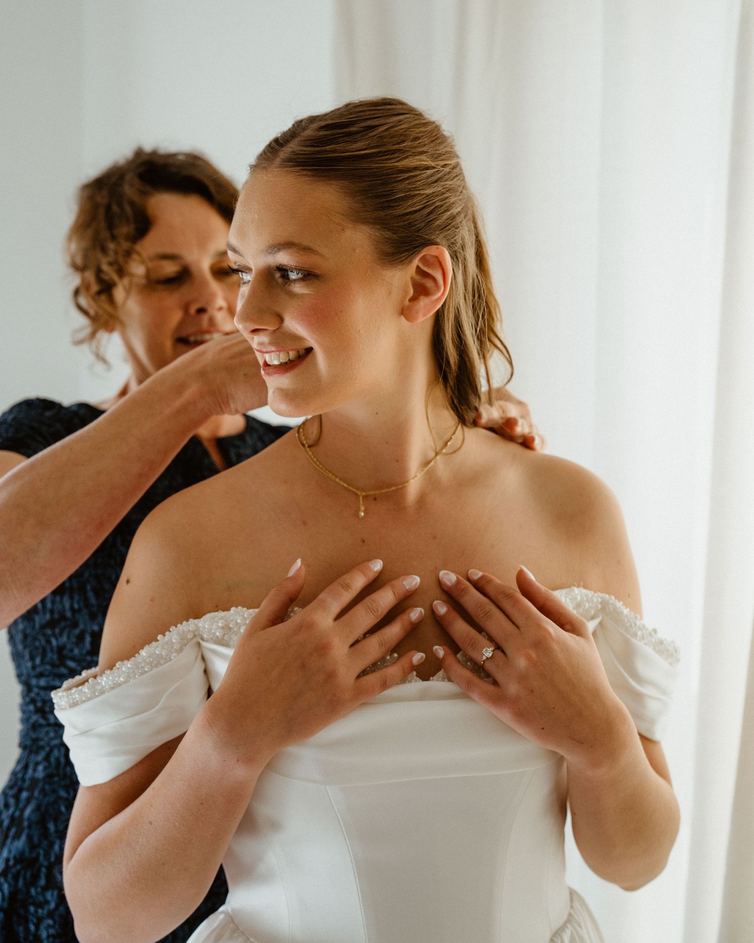 Bride in white off-shoulder gown smiling as woman helps put on gold necklace during wedding preparation.