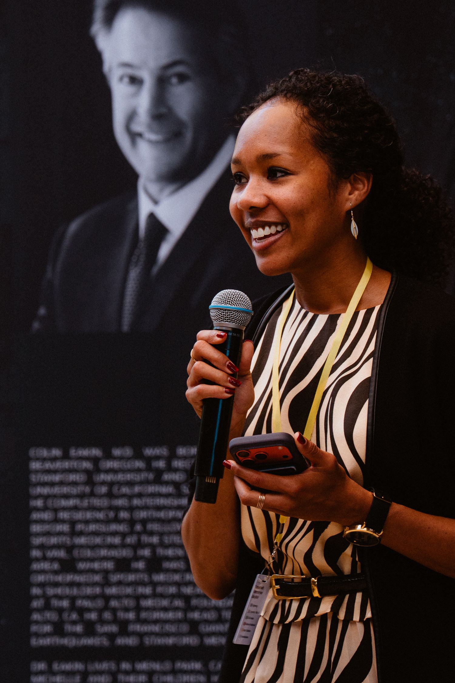 A smiling woman in a zebra-print top holds a microphone and phone while speaking at an event.