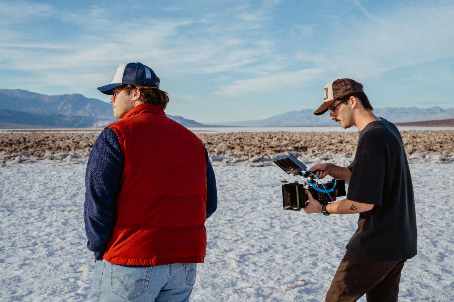 Two men filming on a white salt flat desert landscape with mountains visible in the background.