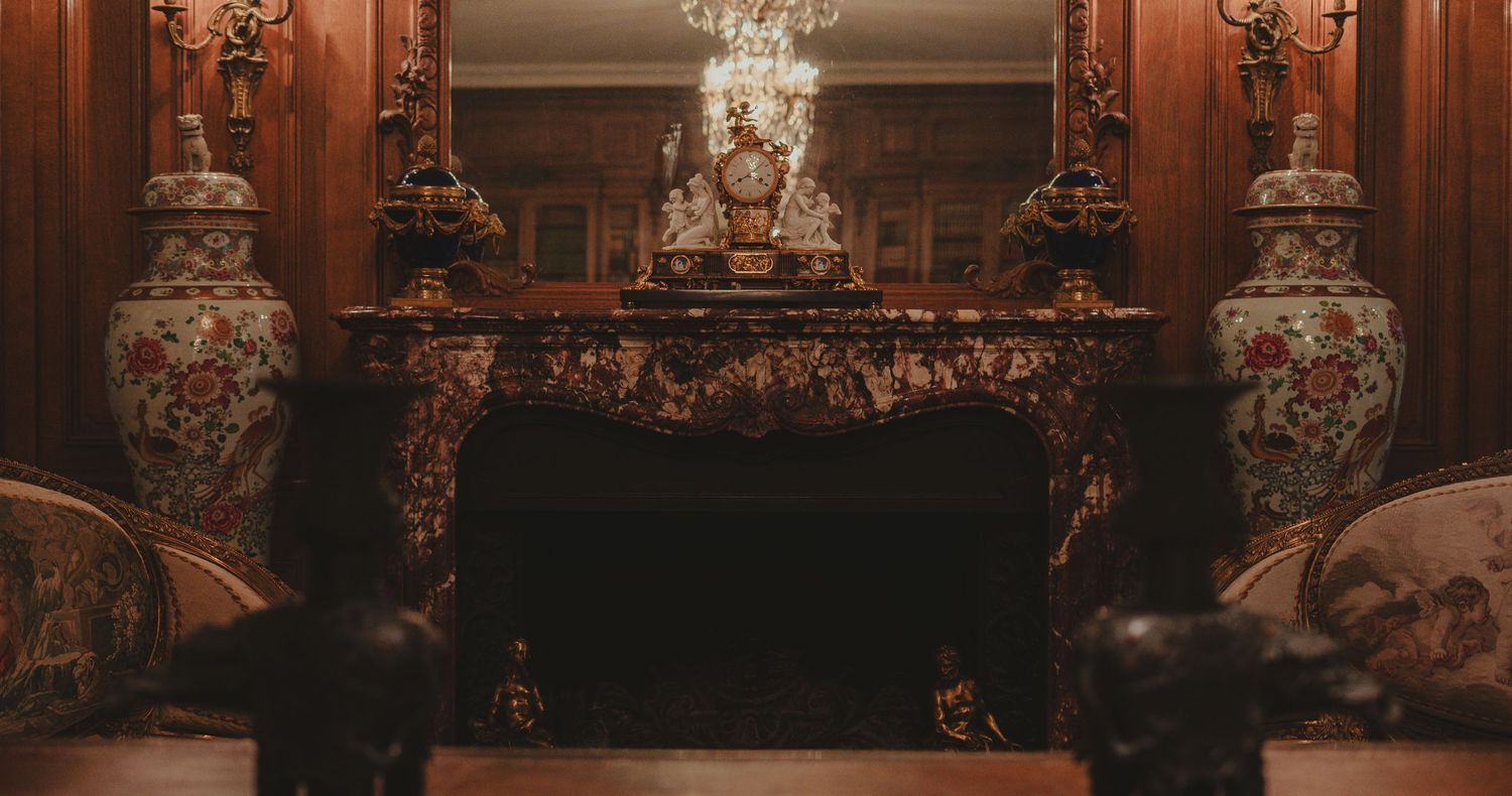 An ornate marble fireplace flanked by decorative vases beneath a gilded mirror in a luxurious room.
