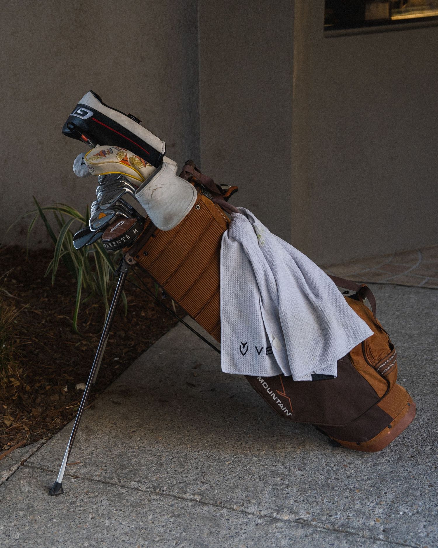 Brown leather golf stand bag with clubs and white towel leaning against concrete wall on sidewalk.