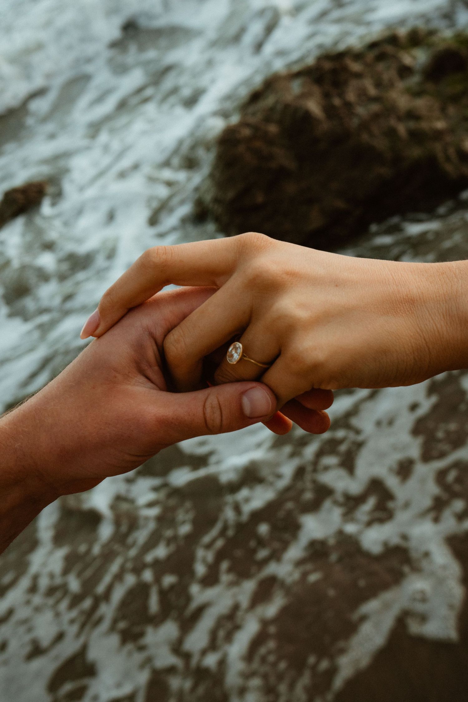 Two hands gently touching, one adorned with a diamond engagement ring, with ocean waves and rocks in the background.