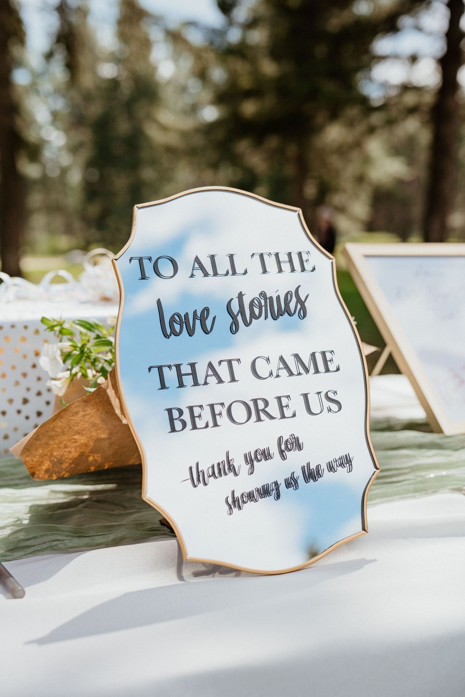 Elegant mirrored wedding sign reading To All The Love Stories That Came Before Us at outdoor reception.