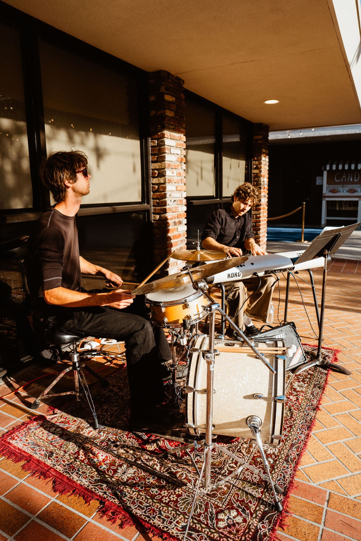 Two musicians playing drums outdoors on a patio with brick pillars and a decorative rug.