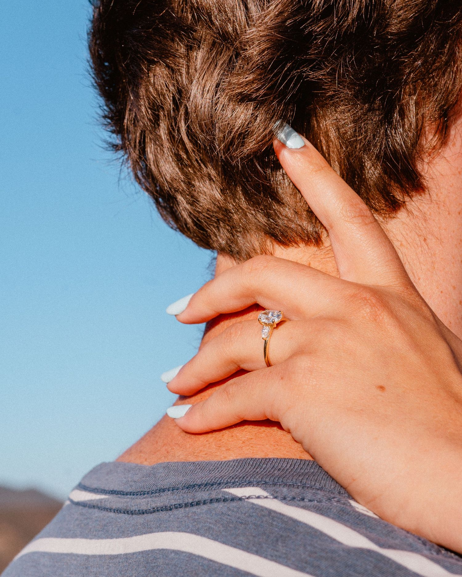 Womans hand with diamond engagement ring and blue nails resting on a mans shoulder against blue sky.