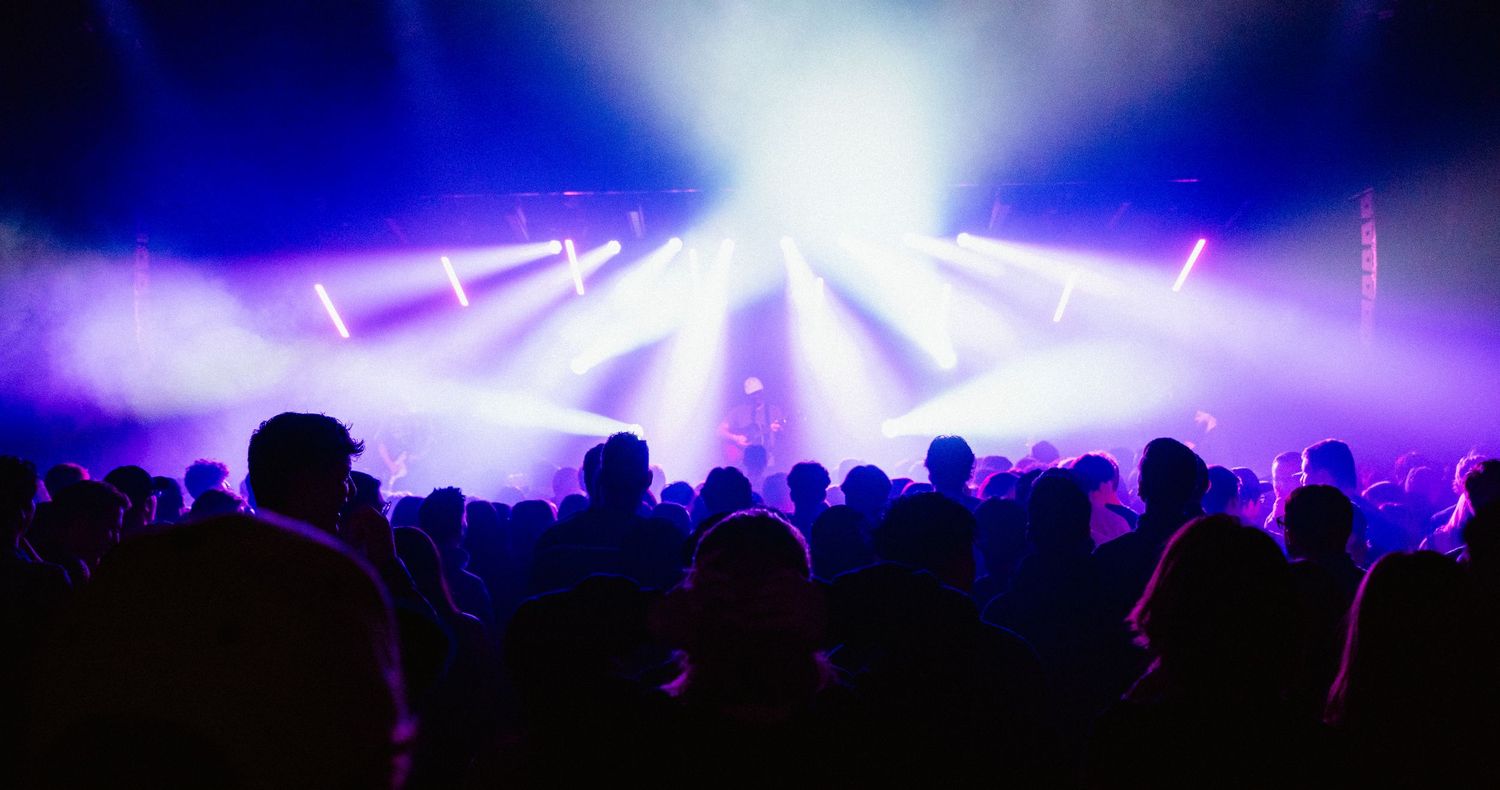 Silhouetted concert crowd bathed in purple and blue stage lights at an indoor music event.