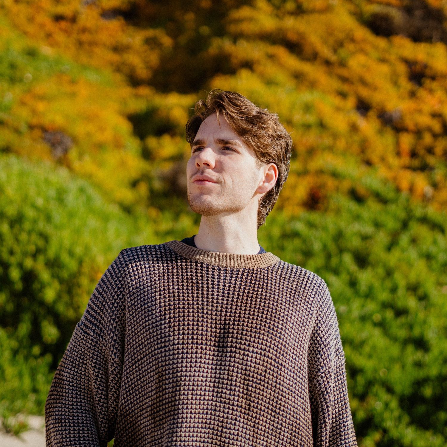 Young man in a knit sweater looking up with colorful autumn foliage in background.