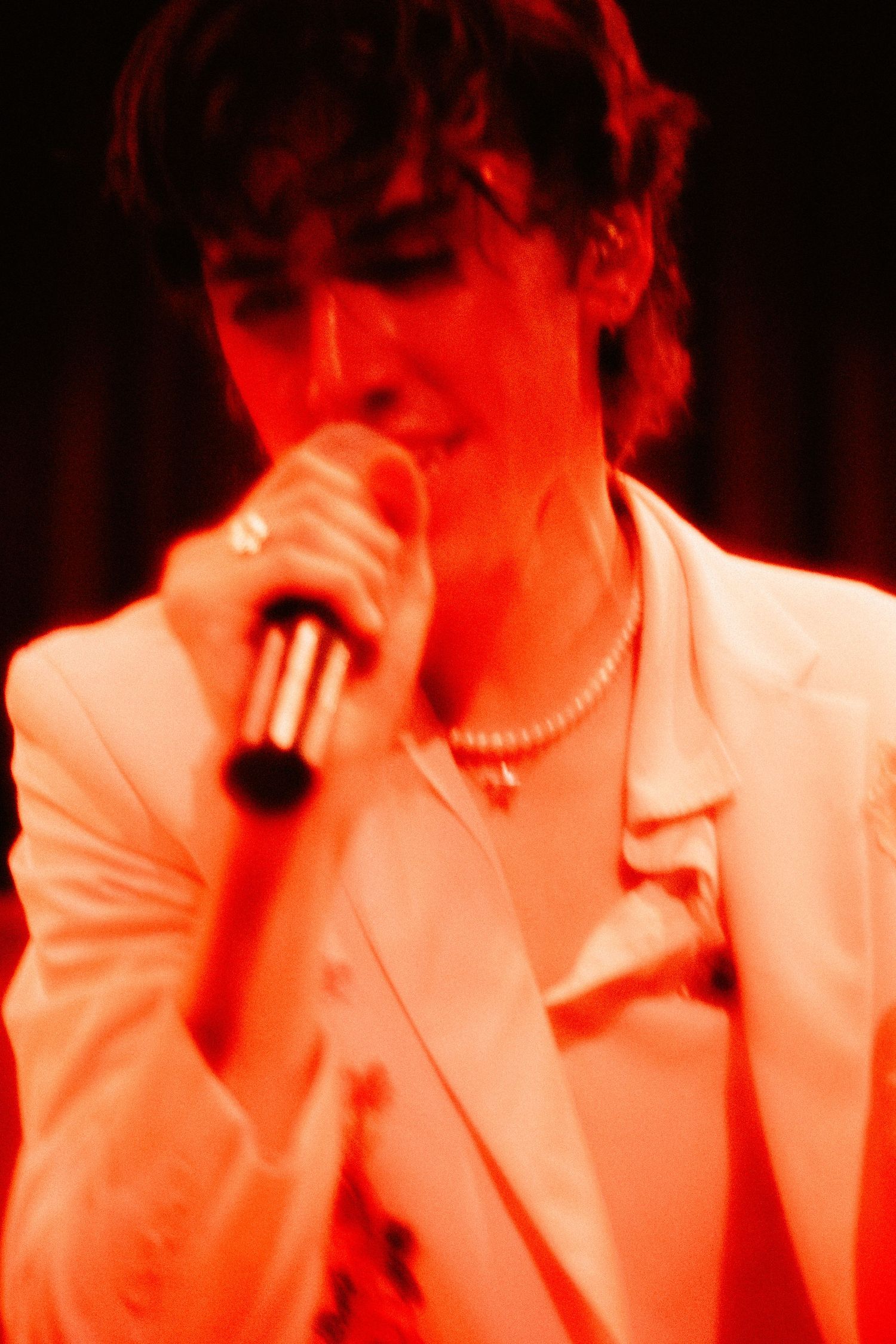 Young male vocalist in white blazer singing into microphone under red stage lighting.