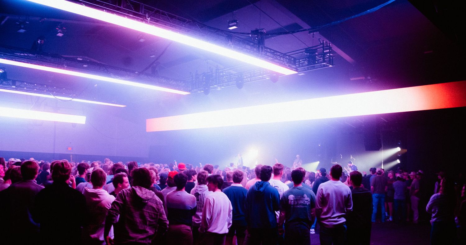 Large crowd gathered under vibrant purple and blue neon lights at an indoor concert venue.