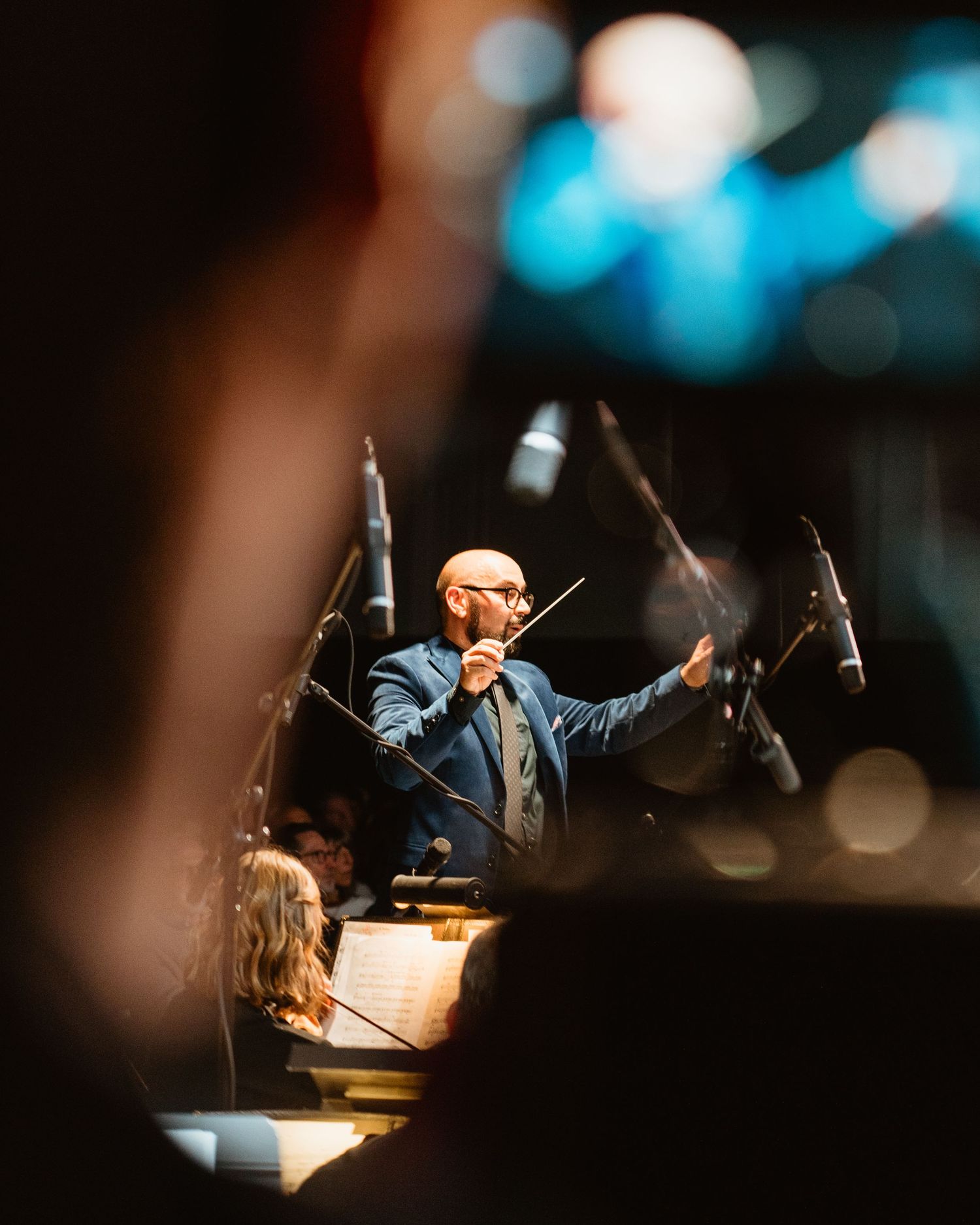 Orchestra conductor leading a symphony performance, holding a baton with microphones and musicians visible in background.