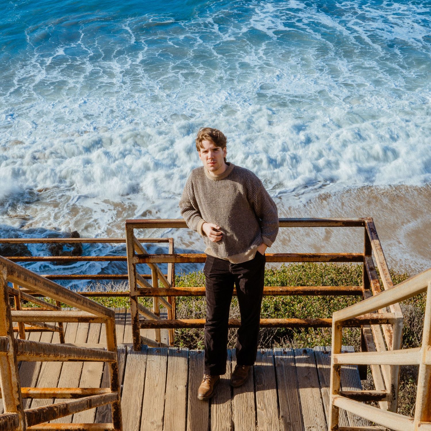 Young man standing on wooden stairs overlooking a turquoise ocean on a sunny day.