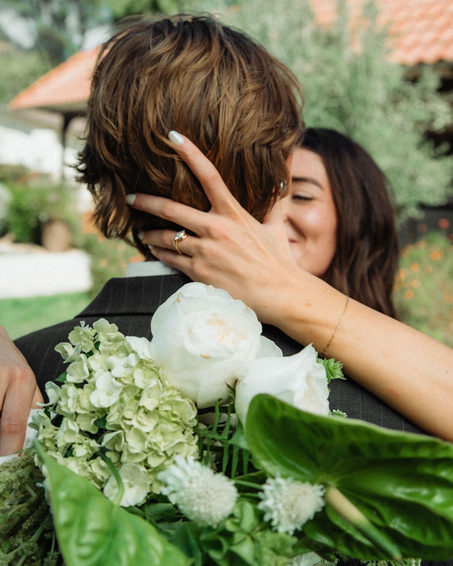 Bride embracing groom while holding white and green floral bouquet with engagement ring visible.