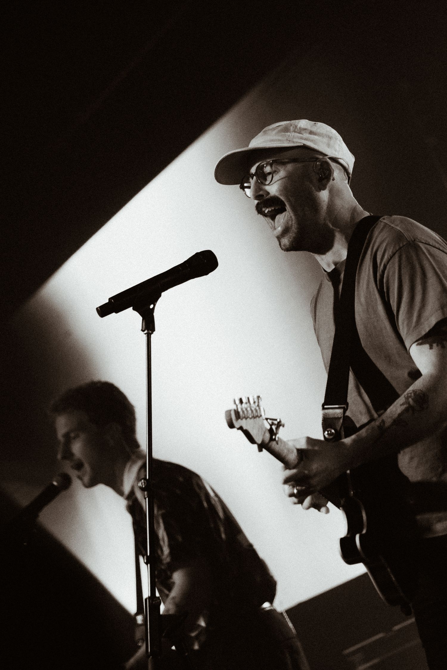 Black and white photo of a guitarist singing passionately on stage with a cap and glasses.
