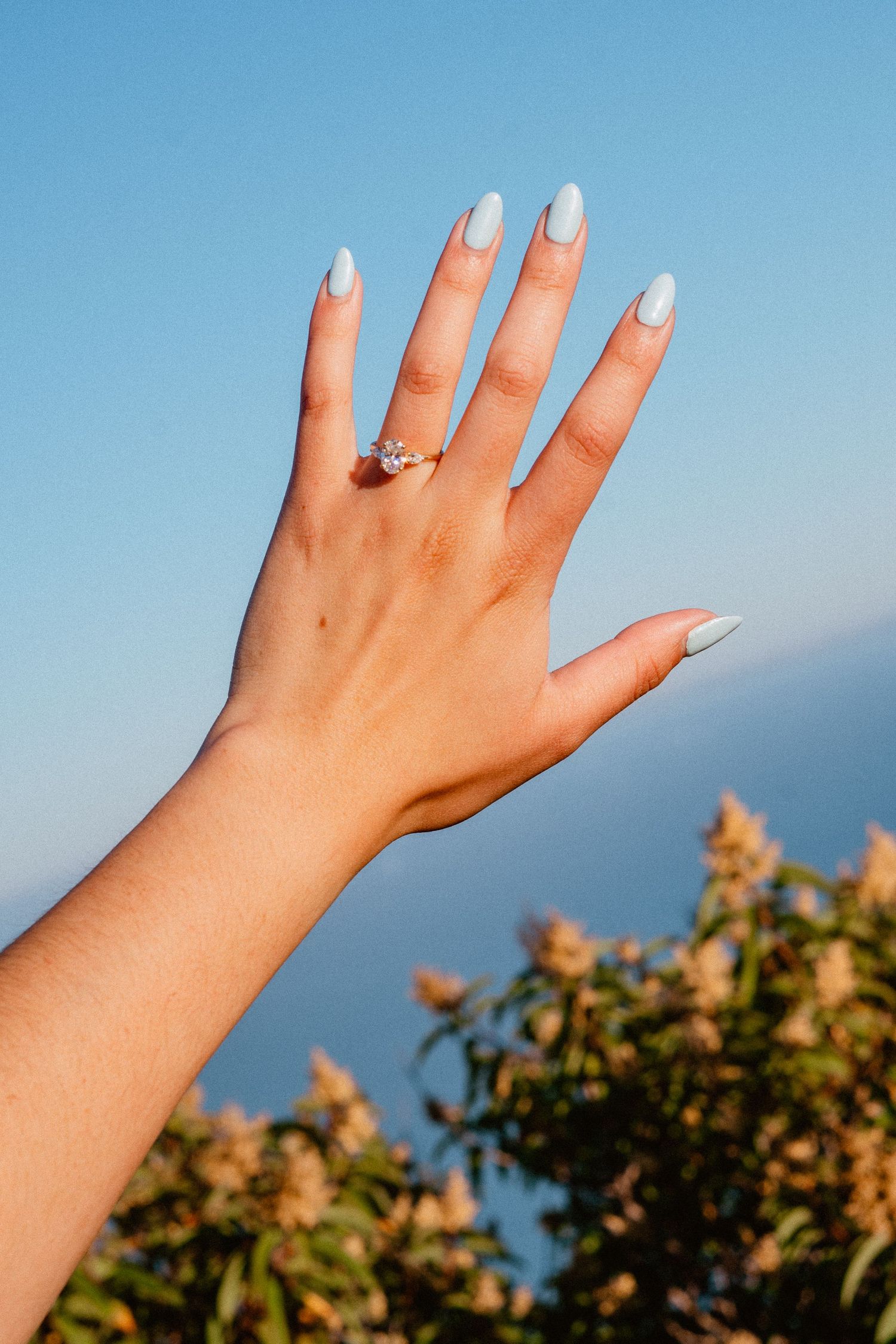 Close-up of a womans hand displaying a diamond engagement ring with light blue nails against blue sky.