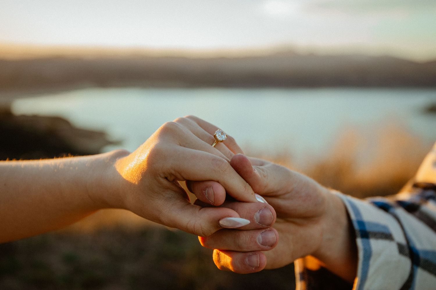 Close-up of couple holding hands with engagement ring at golden hour by a lake.