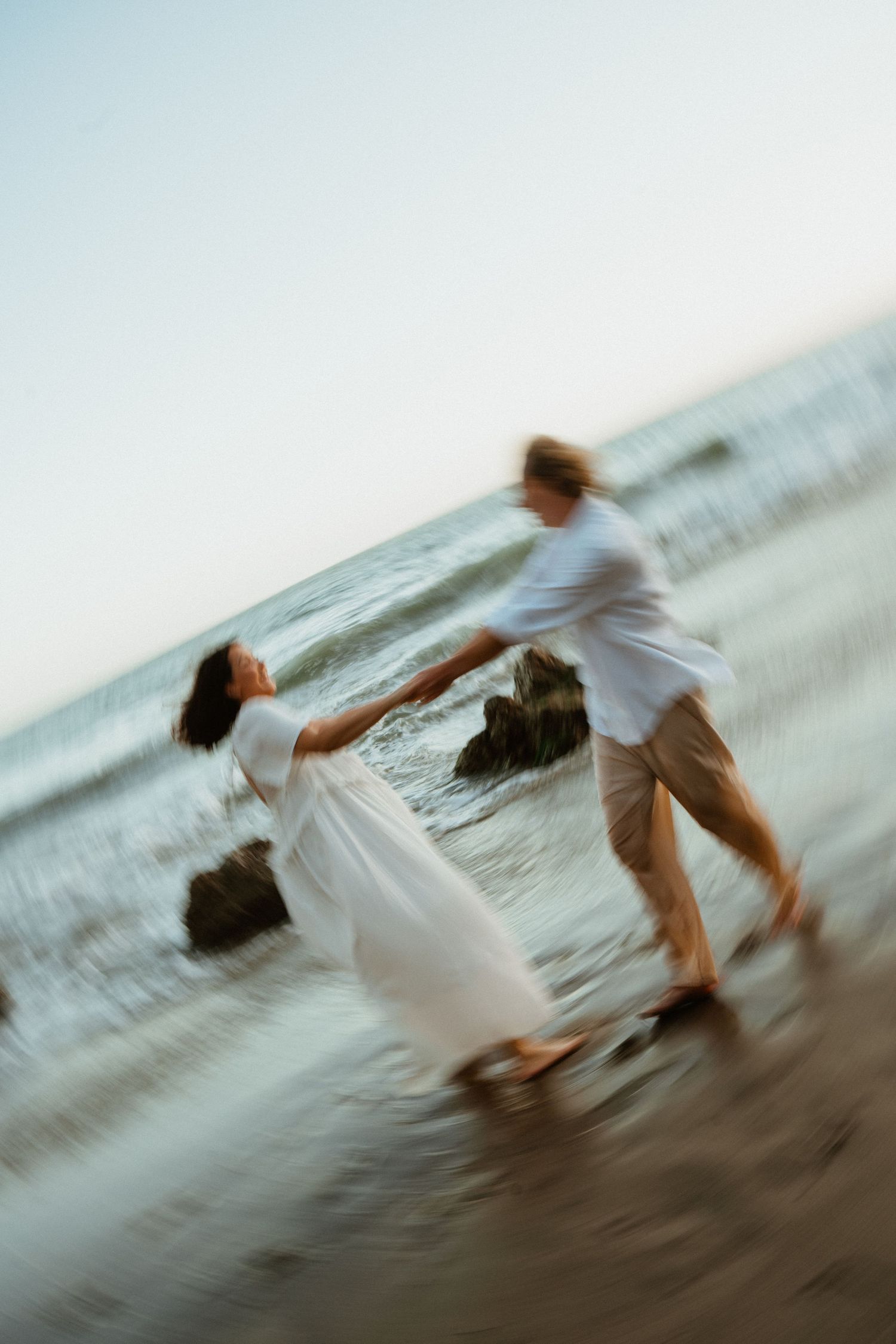 Blurred motion photo of a romantic couple holding hands and dancing on a beach at sunset near ocean waves.