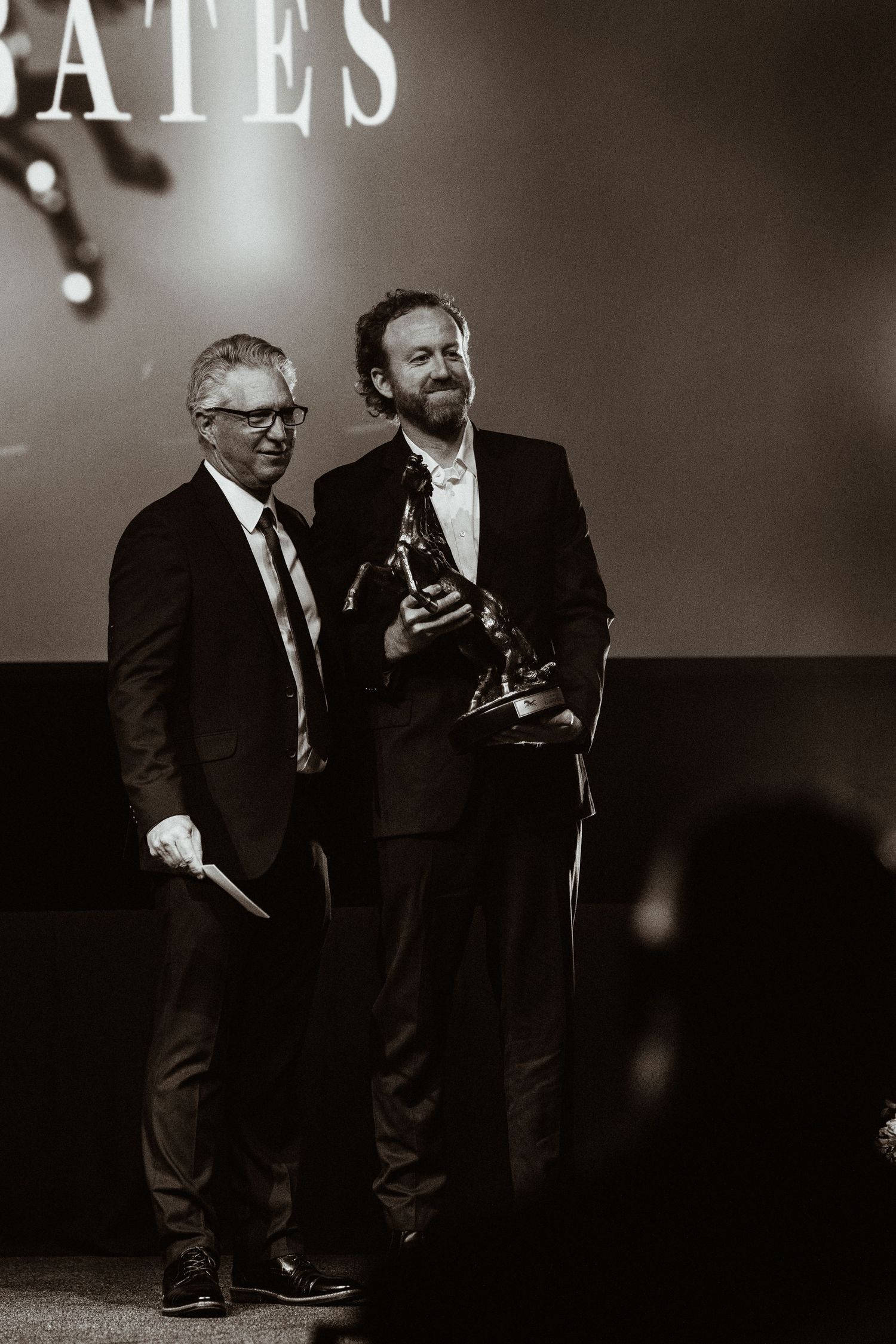 Two men in suits smile on stage, one holding a trophy award, in black and white photography.