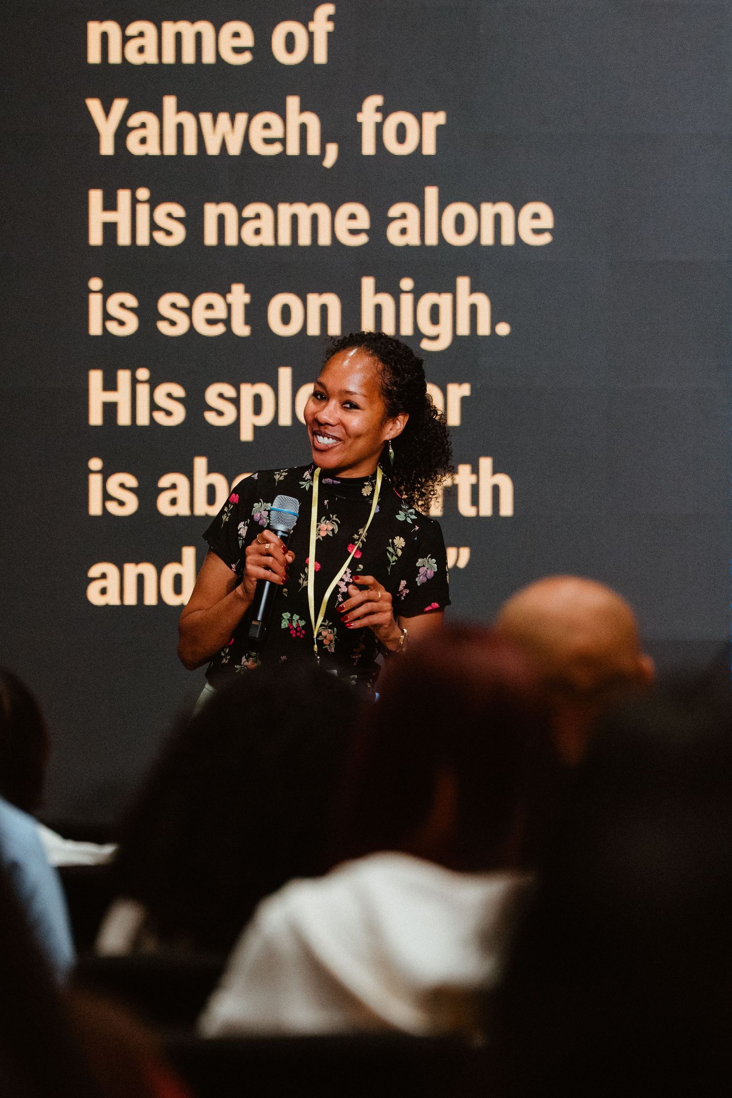 Woman smiling and holding microphone speaks at religious event with Psalm scripture projected on screen behind her.