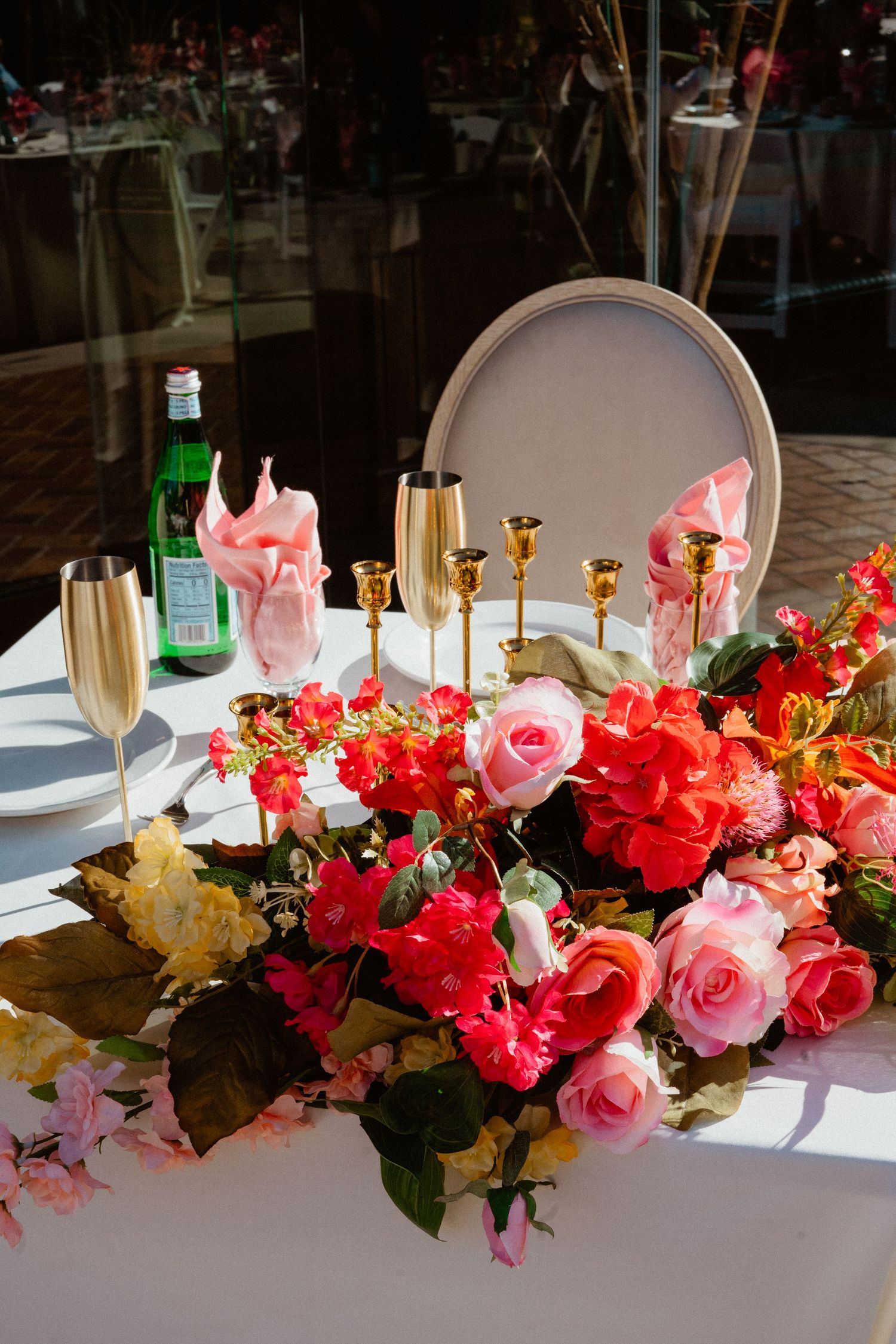 Wedding sweetheart table with gold candle holders, champagne flutes, lush red and pink floral arrangement, and pink napkins.