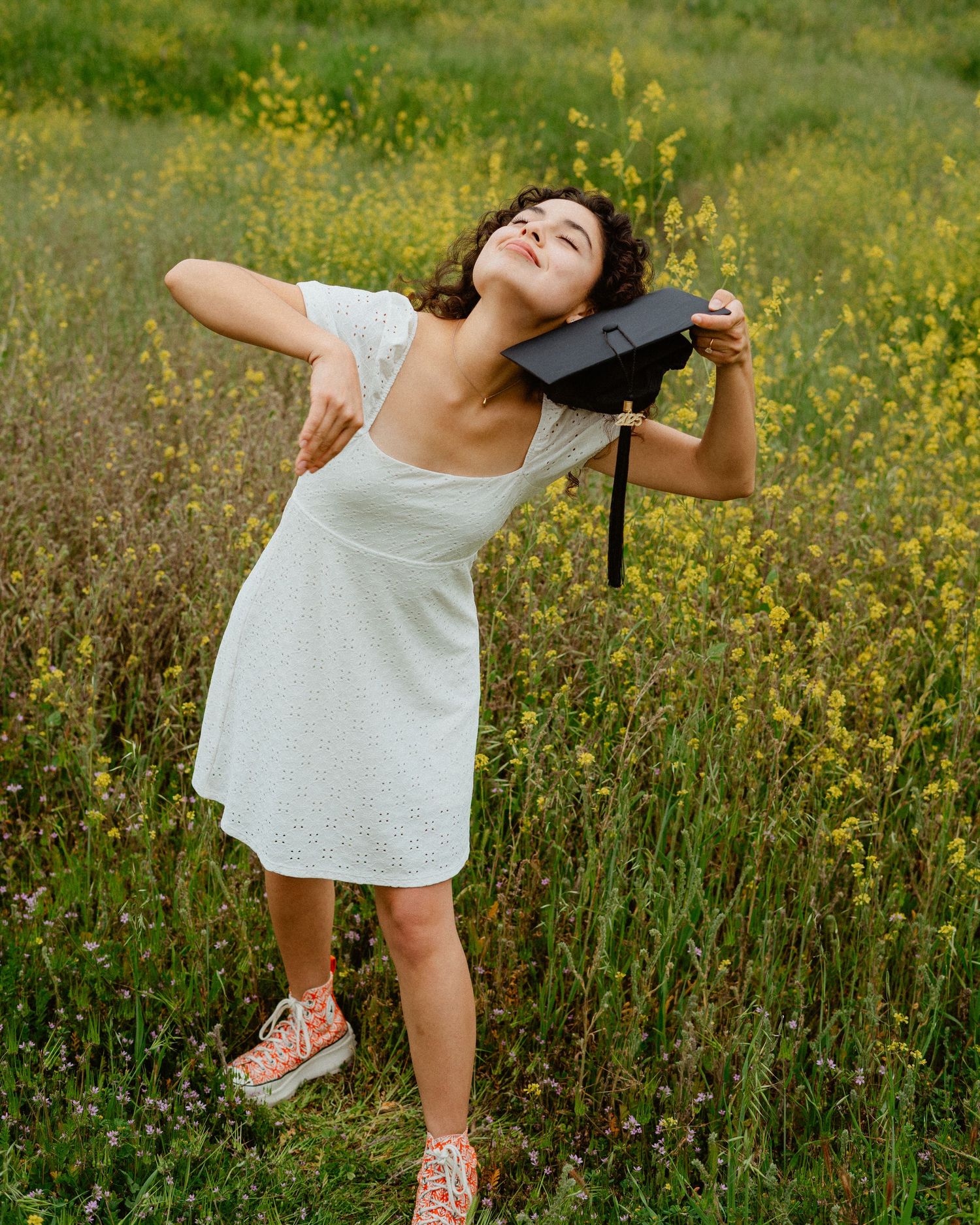 Young woman in white dress playfully holding graduation cap in a yellow wildflower field.