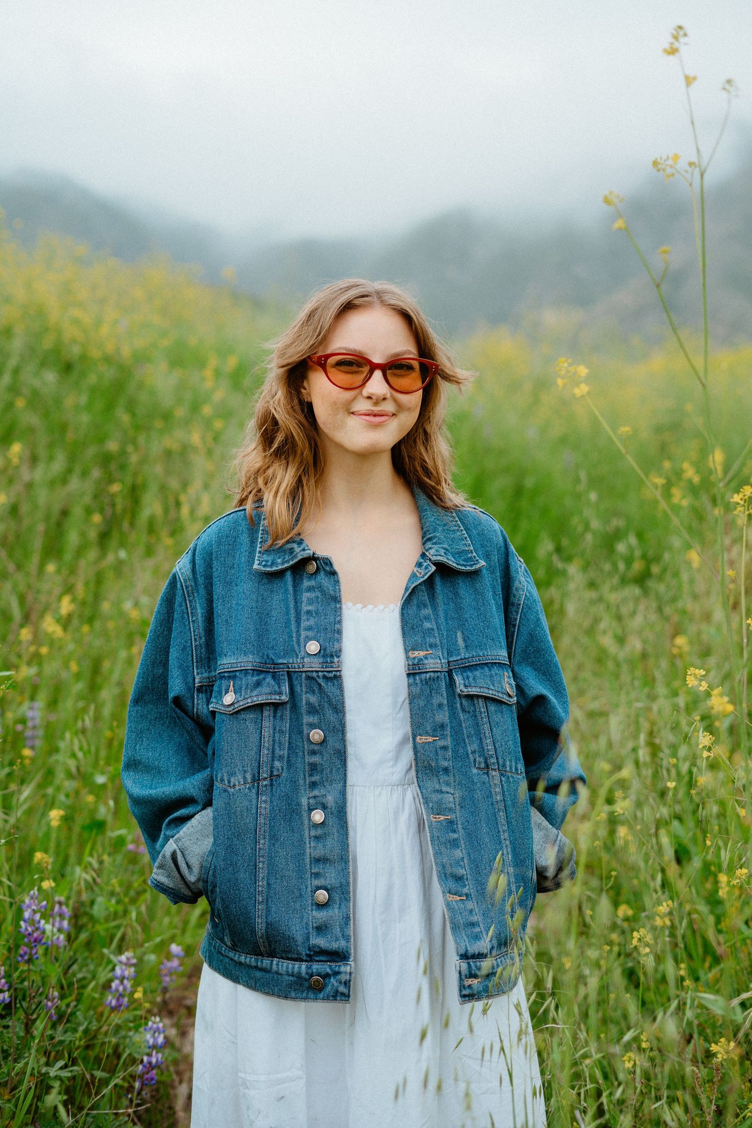 Young woman in oversized denim jacket, white dress, and orange sunglasses poses in a wildflower field.