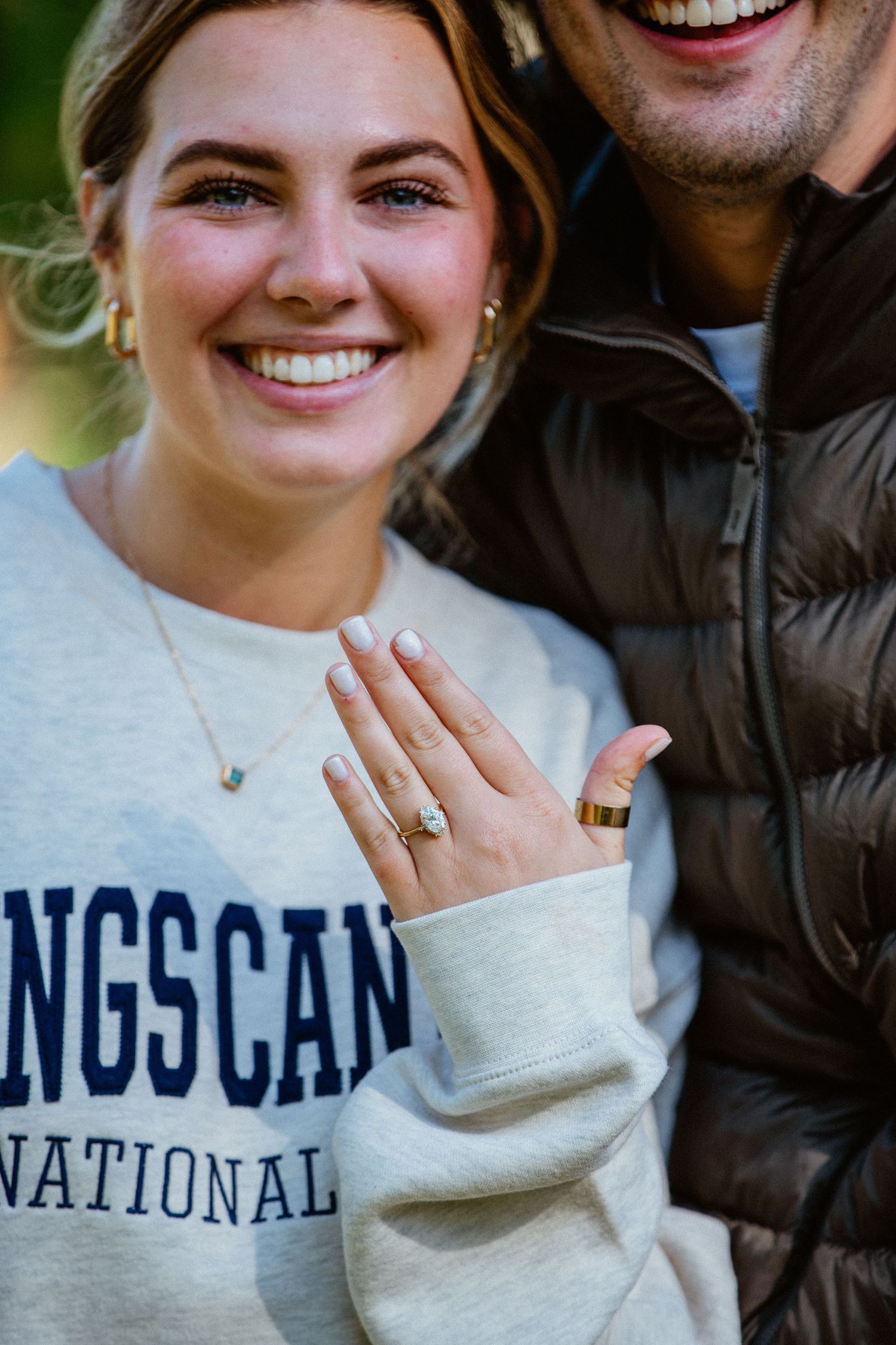 Close-up of smiling woman displaying oval diamond engagement ring on her hand.