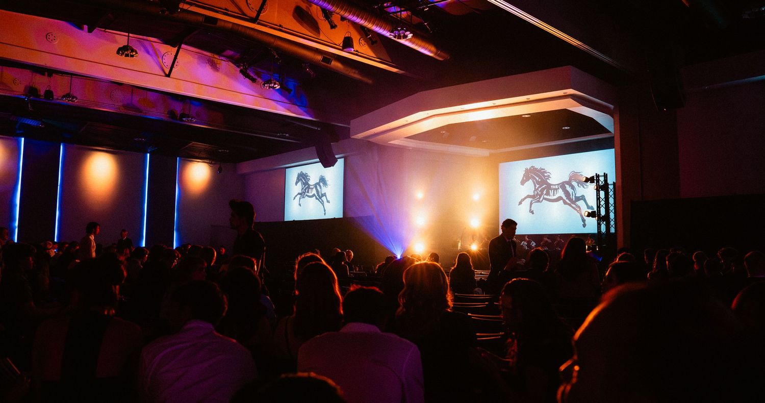 Audience watching a presentation with horse silhouette projections on dual screens in a dimly lit event venue.