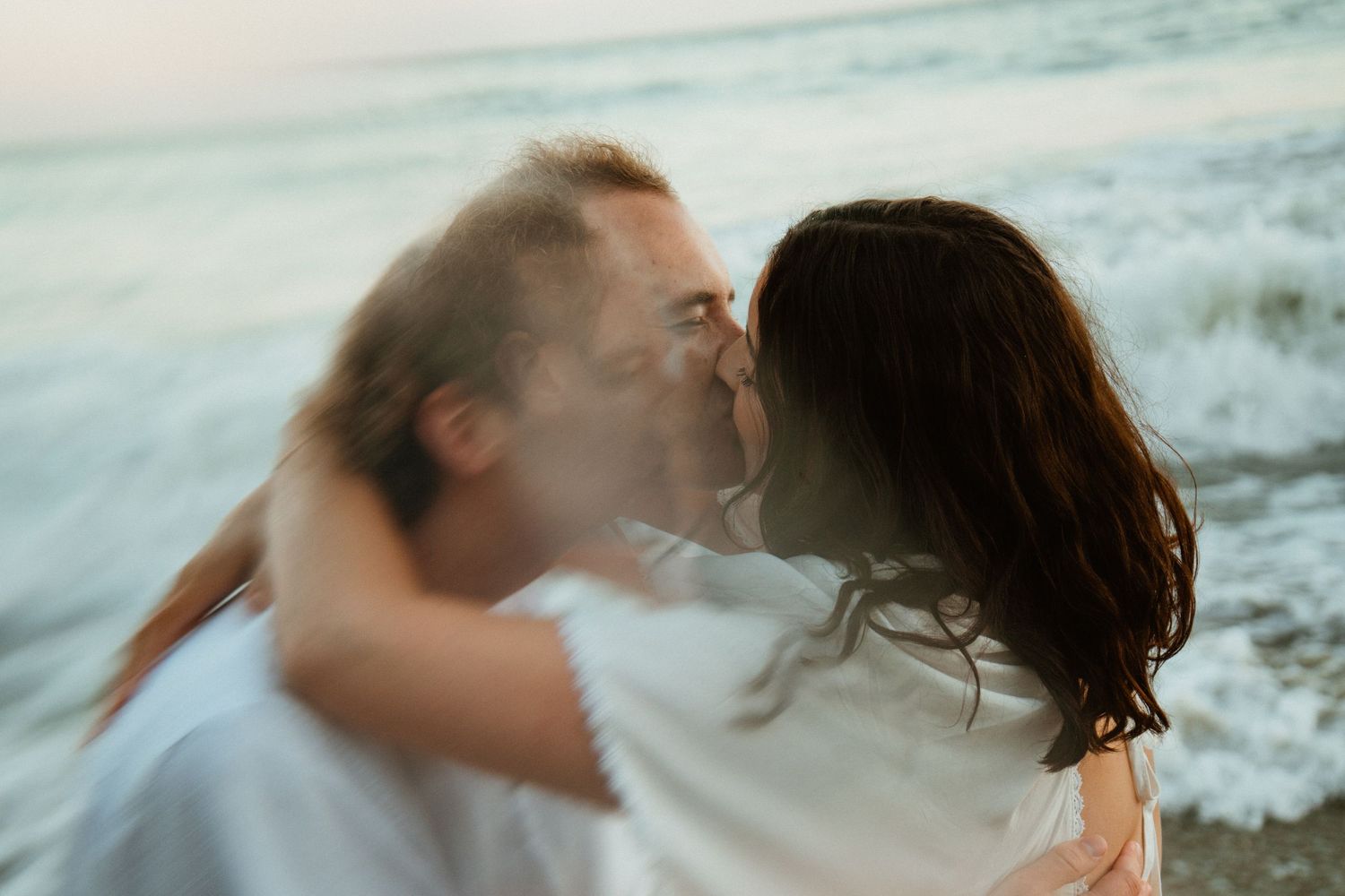 A couple sharing a romantic kiss on a beach with ocean waves in the background, captured with motion blur.