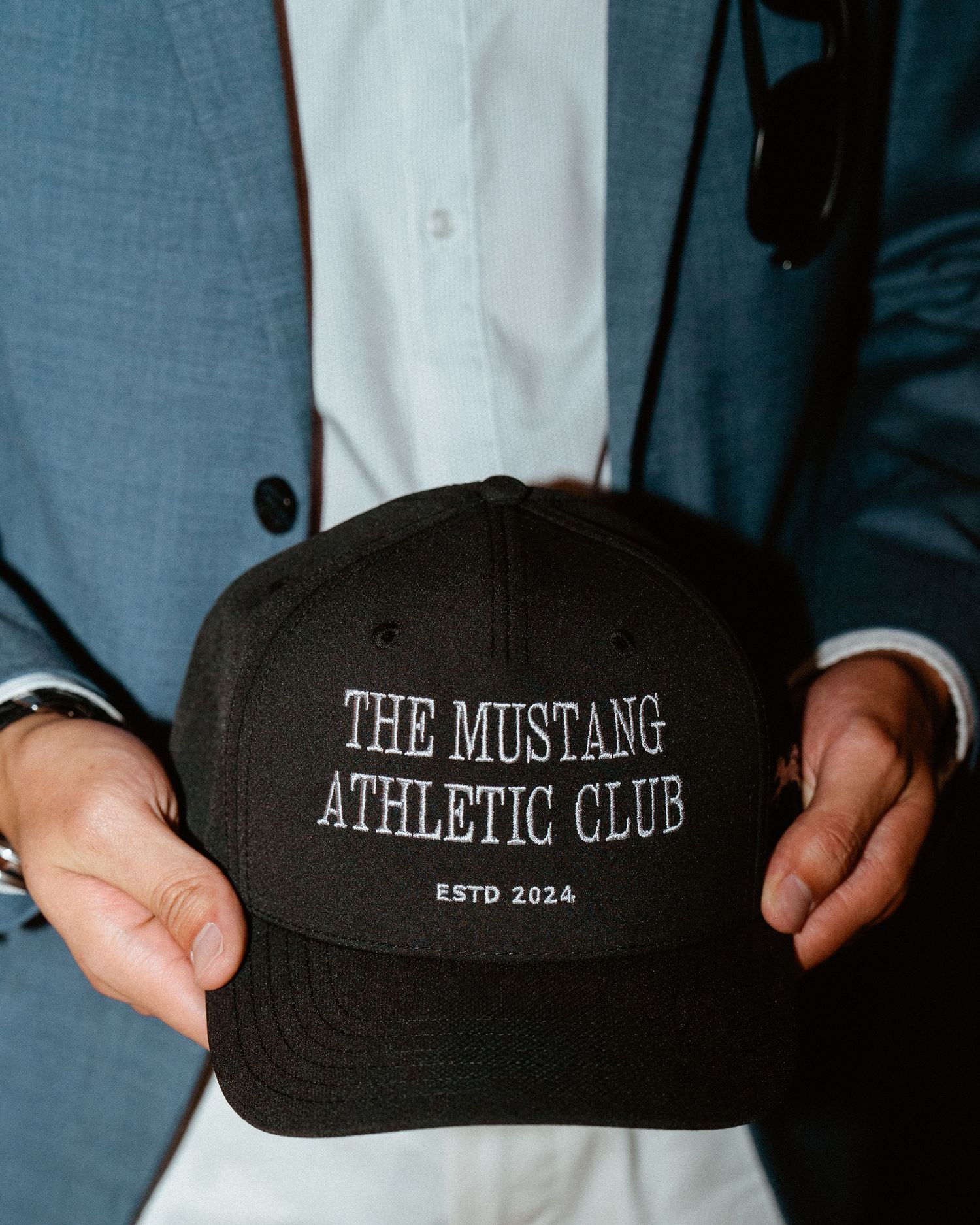 Person holding a black snapback cap embroidered with The Mustang Athletic Club Est 2024 logo.