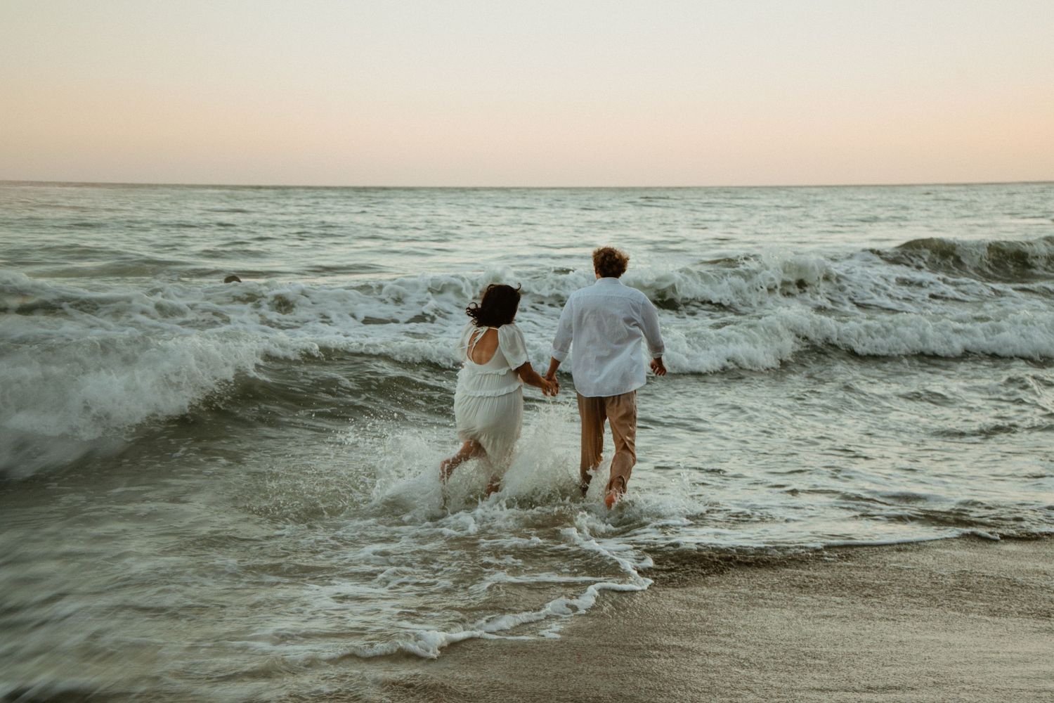 Couple holding hands running into ocean waves at sunset on a sandy beach wearing white outfits.