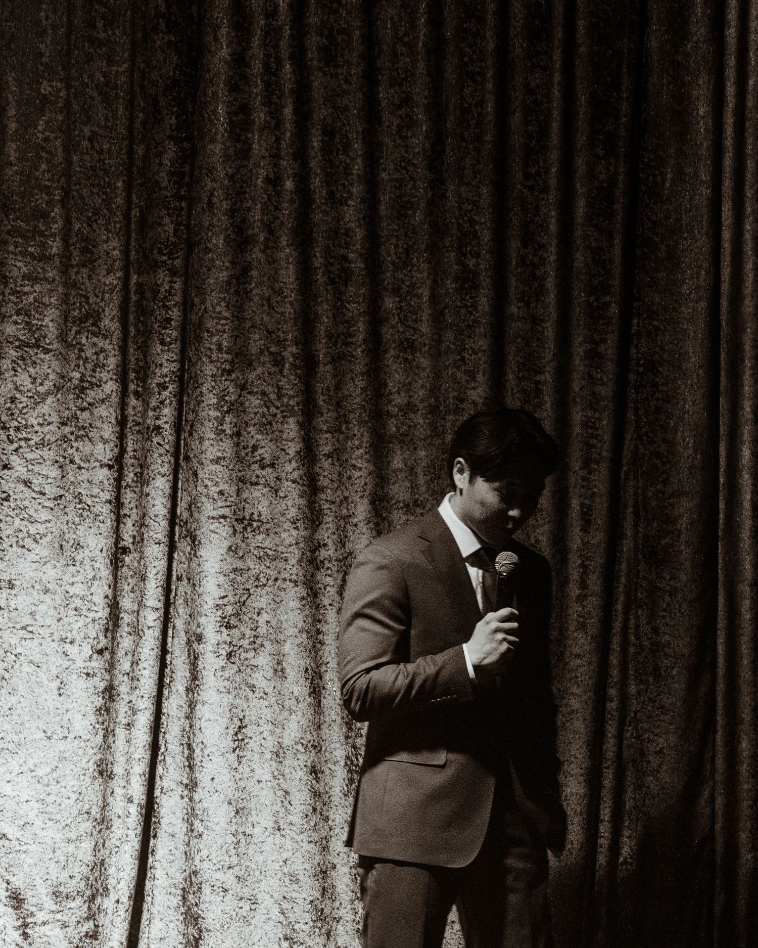 Man in suit holding microphone standing before velvet curtain on stage.