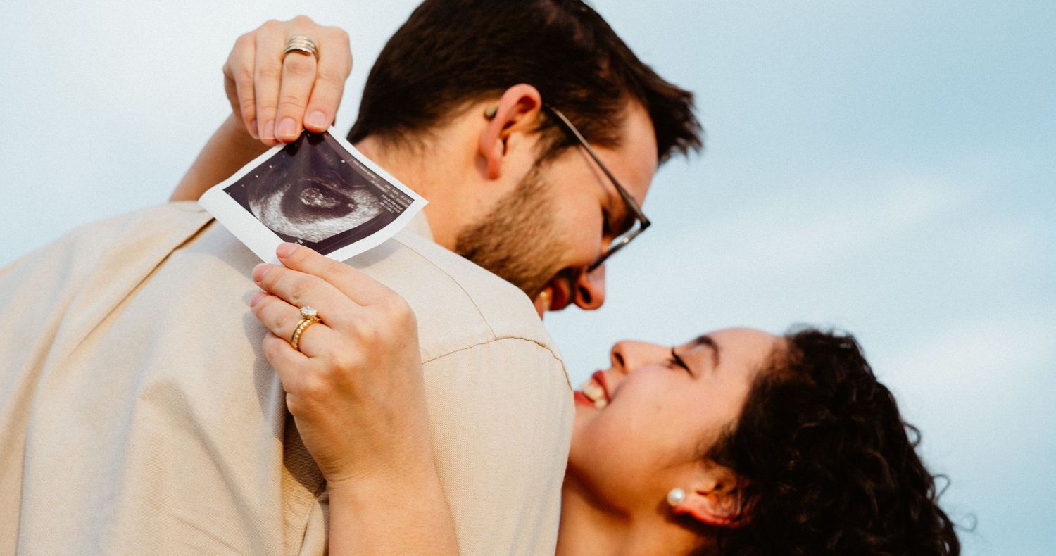 Joyful couple laughing together while holding ultrasound image for outdoor pregnancy reveal photo.