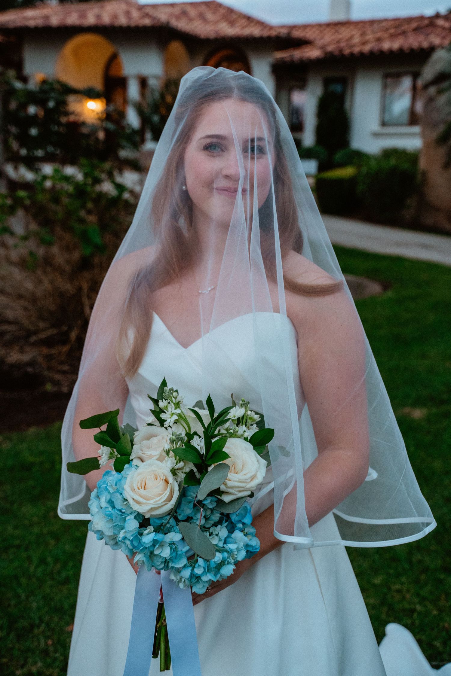 Elegant bride wearing sheer veil and white strapless dress holds blue and white floral bouquet.