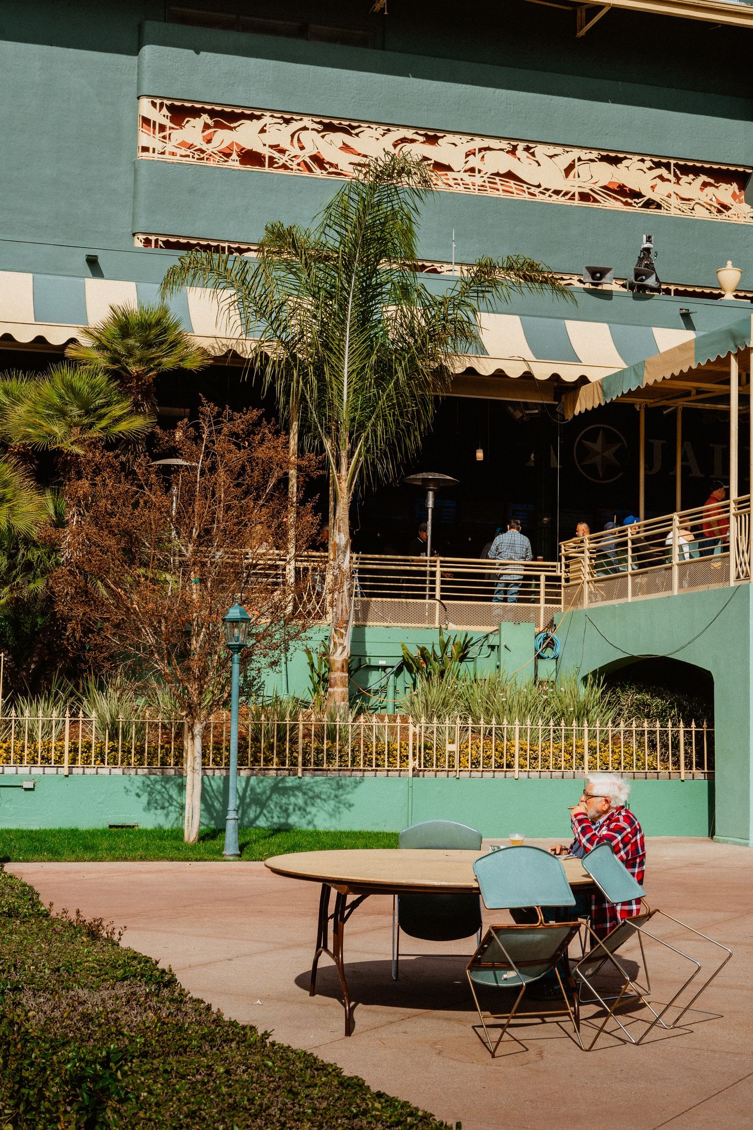 Person in cowboy hat sitting at outdoor table near a teal Art Deco racetrack building.