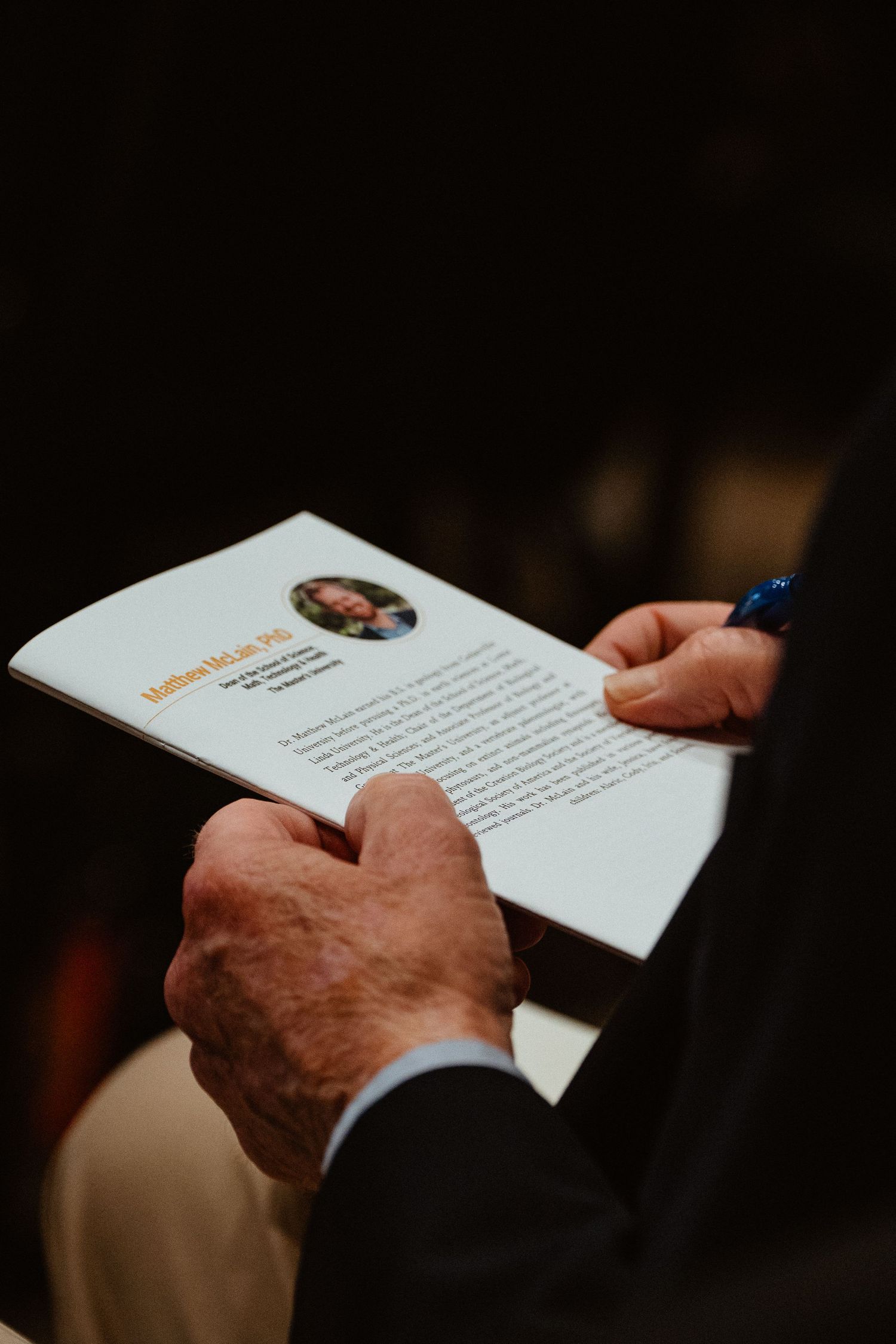 Elderly man holding a program booklet with a photo and pen at a formal event.