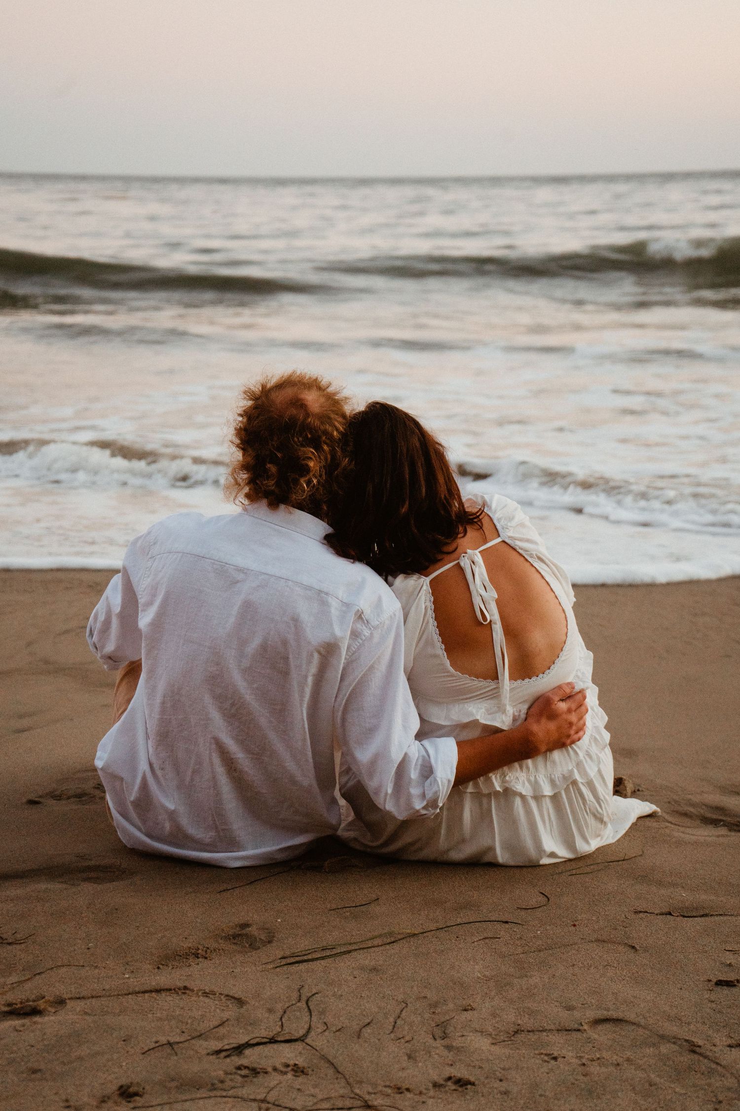 Couple in white outfits sitting together on sandy beach, embracing while watching ocean waves at sunset.