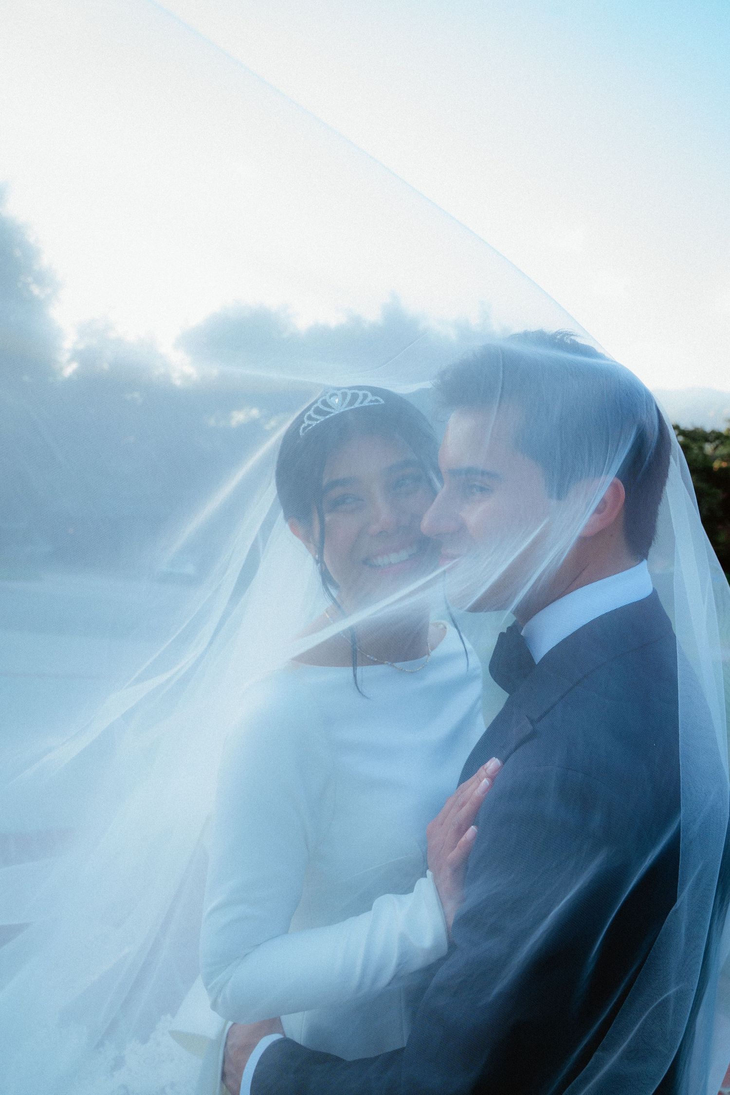 Romantic couple embracing under sheer wedding veil, smiling together in soft natural light outdoors.
