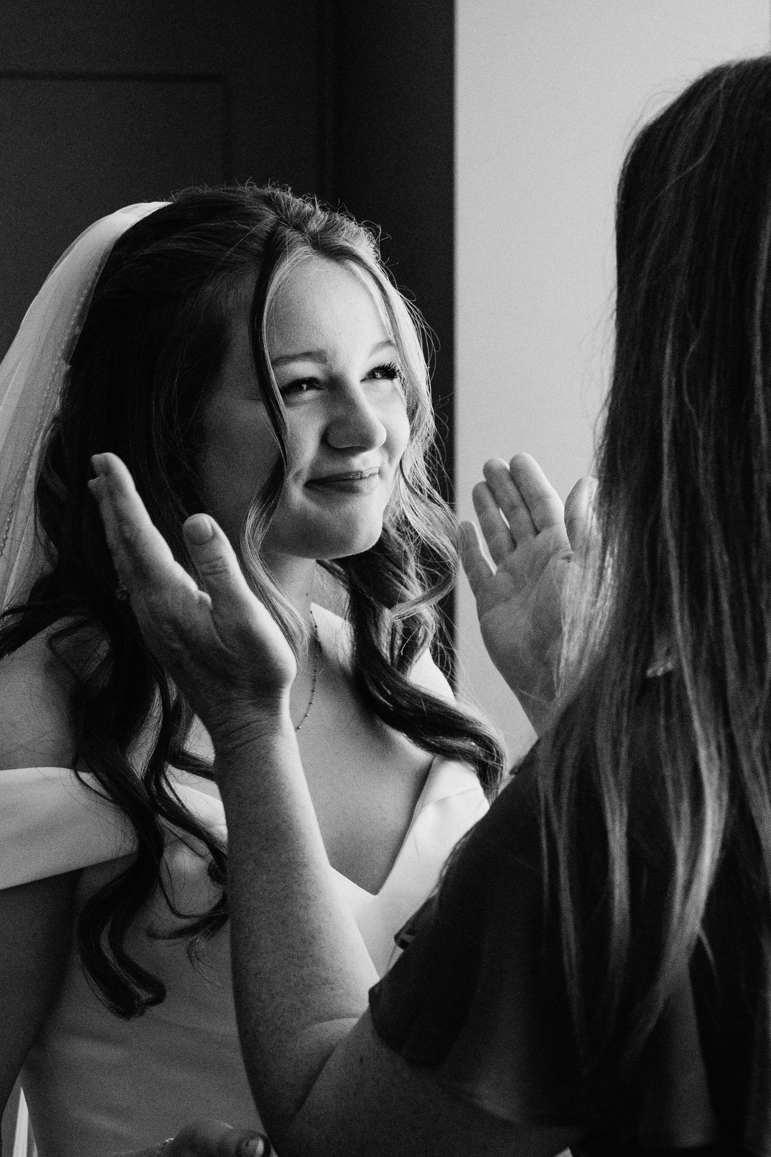 Black and white photo of a smiling bride in veil having makeup applied on her wedding day.